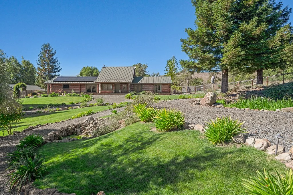 A spacious yard with well-maintained lawns, decorative plants, and large trees. There is a brown house with a metal roof in the background, and the sky is clear and blue.