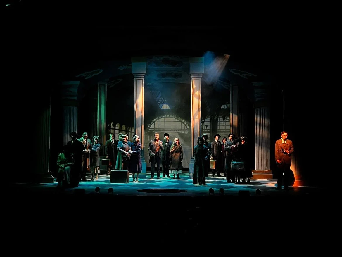 Theater stage with actors in period costumes, standing in formation, lit by stage lights and a backdrop resembling a train station.