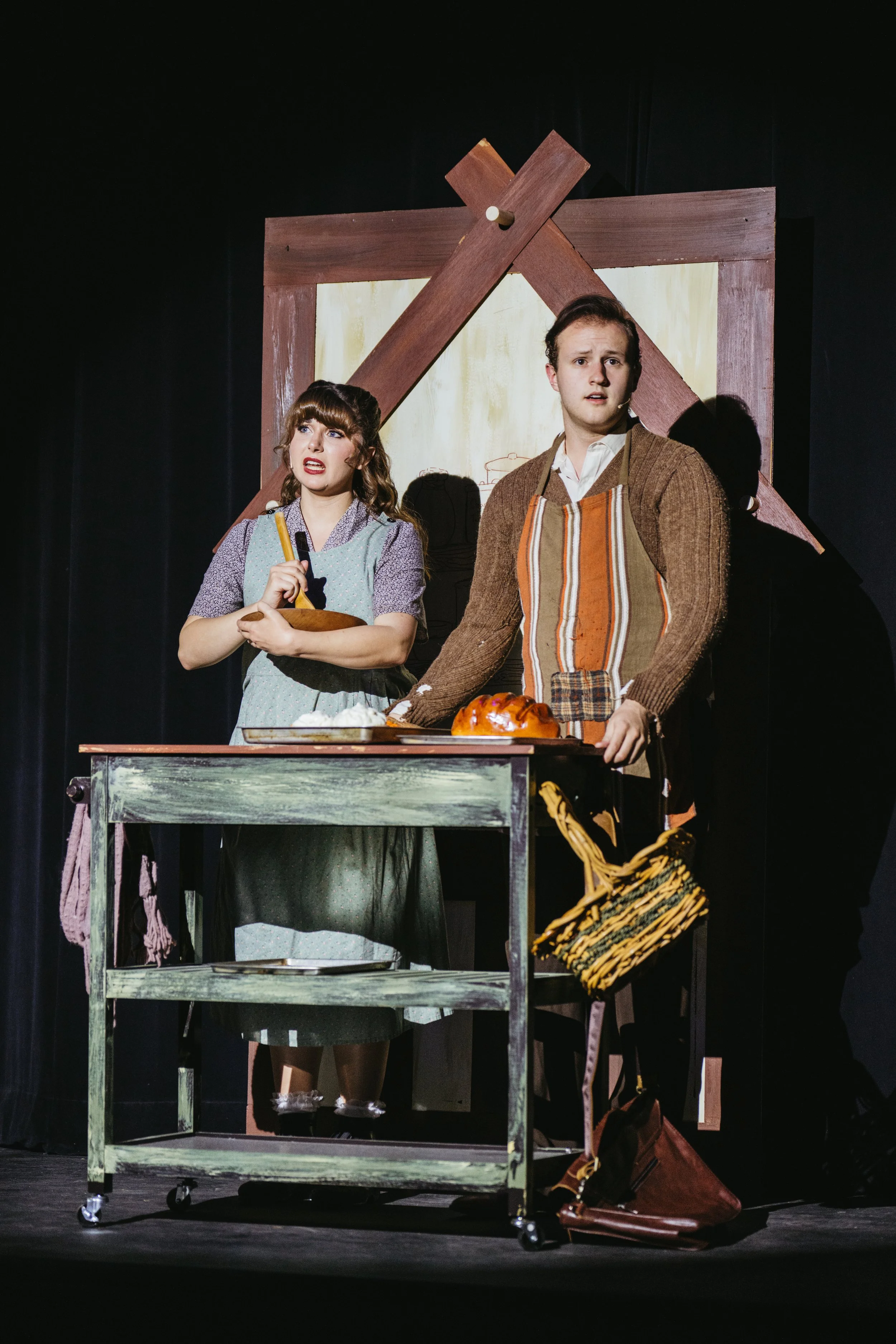 Two actors on stage in a theatrical play, with one holding a loaf of bread and a basket, and the other holding a bowl, in front of a rustic set with painted wooden frame and a large wooden cross.