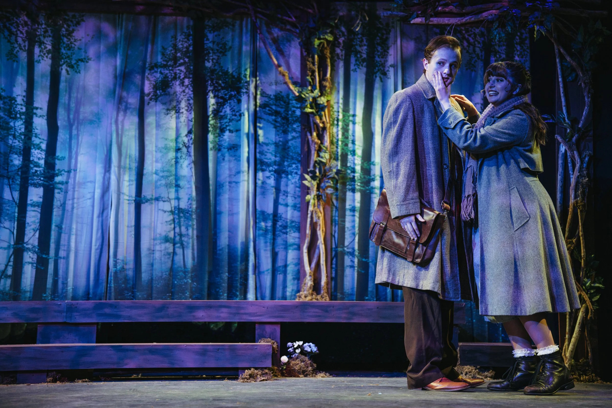 Two actors on stage acting in a play set in a forest, with a backdrop of trees and greenery. The actor on the left has short hair, is wearing a gray coat and carrying a brown bag. The actor on the right has long hair, is wearing a gray coat and boots