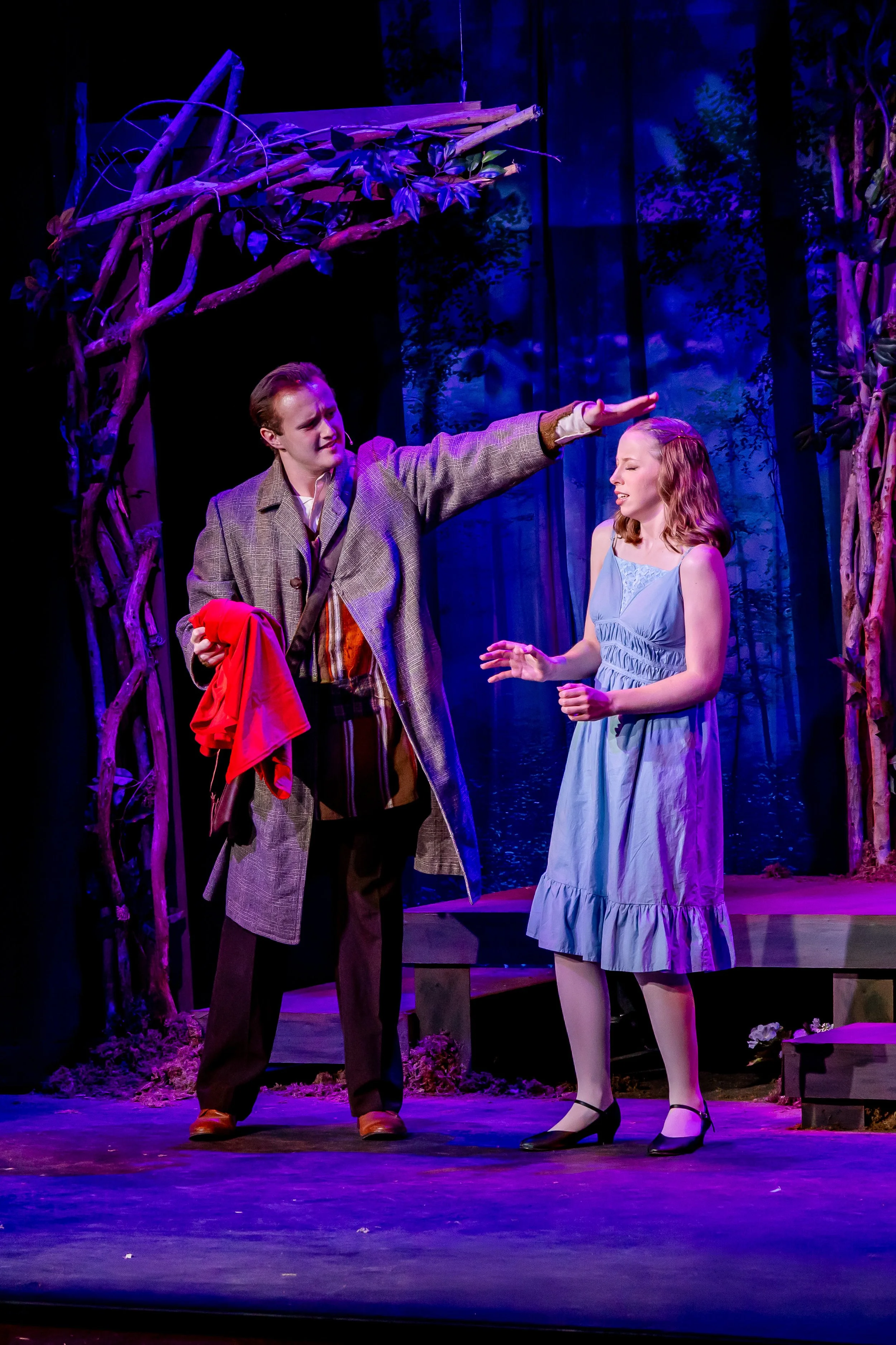 Two actors performing on stage, with the male actor gesturing as if stopping or blocking the female actor, in a scene with a blue-lit forest background and rustic props.