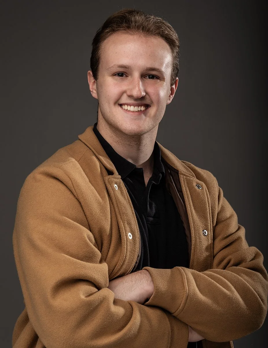 A young man with light skin, short brown hair, and blue eyes, smiling with arms crossed, wearing a tan jacket over a black shirt, against a dark background.