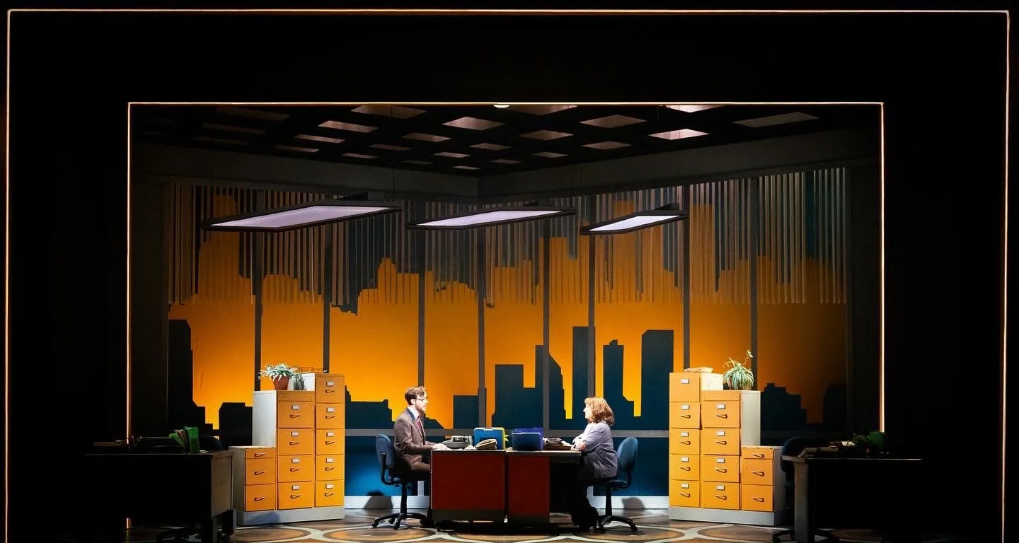 Two people sitting at desks in an office, with a city skyline silhouette background painted on the wall, illuminated by overhead lights.