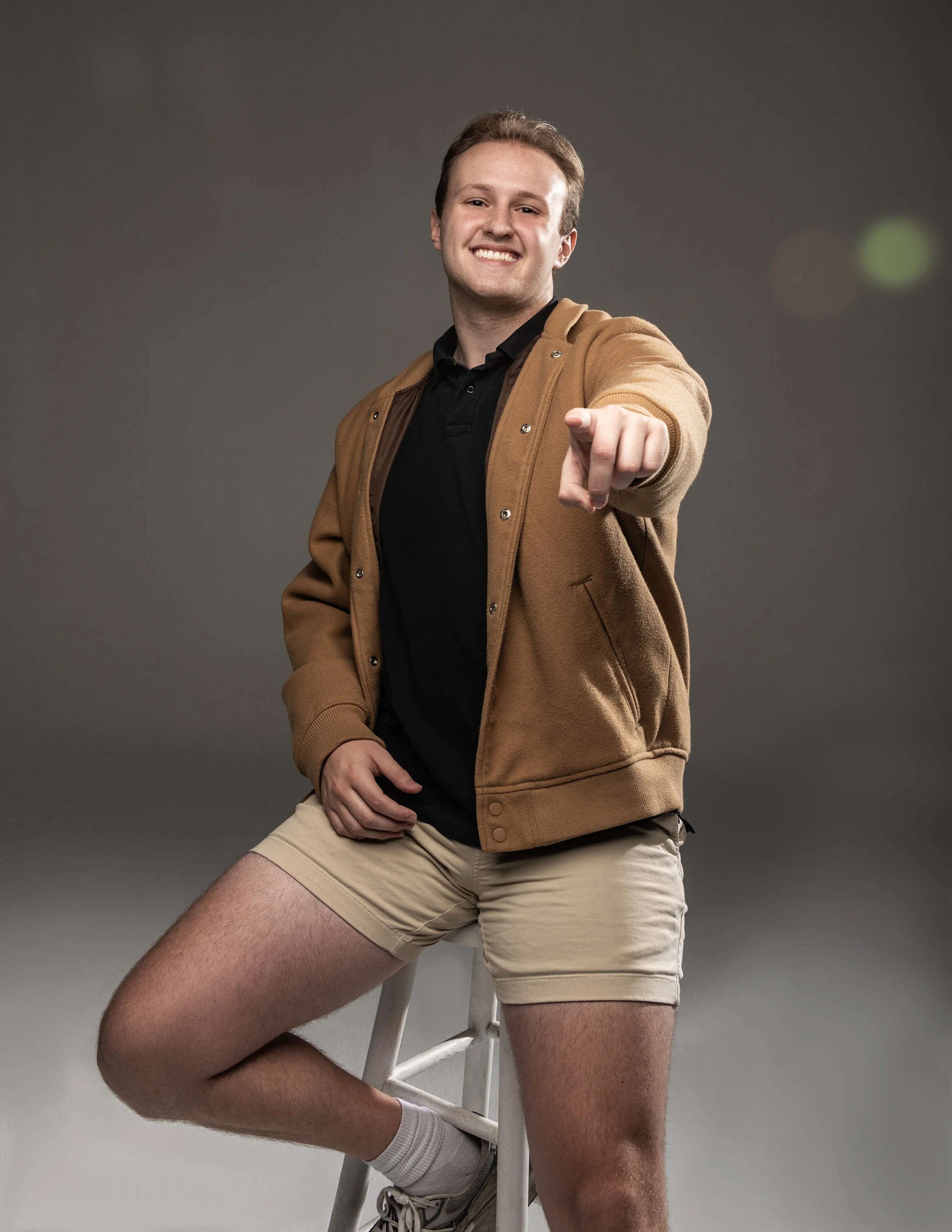 A young man with short brown hair, smiling and pointing at the camera, sitting on a stool in front of a gray background, wearing a brown jacket, black polo shirt, beige shorts, white socks, and gray sneakers.