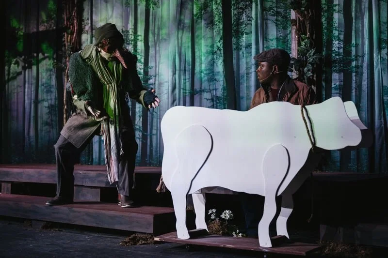 Two actors on a stage with a forest backdrop, one dressed as a witch and the other as a farmer, with a large white cow prop between them.