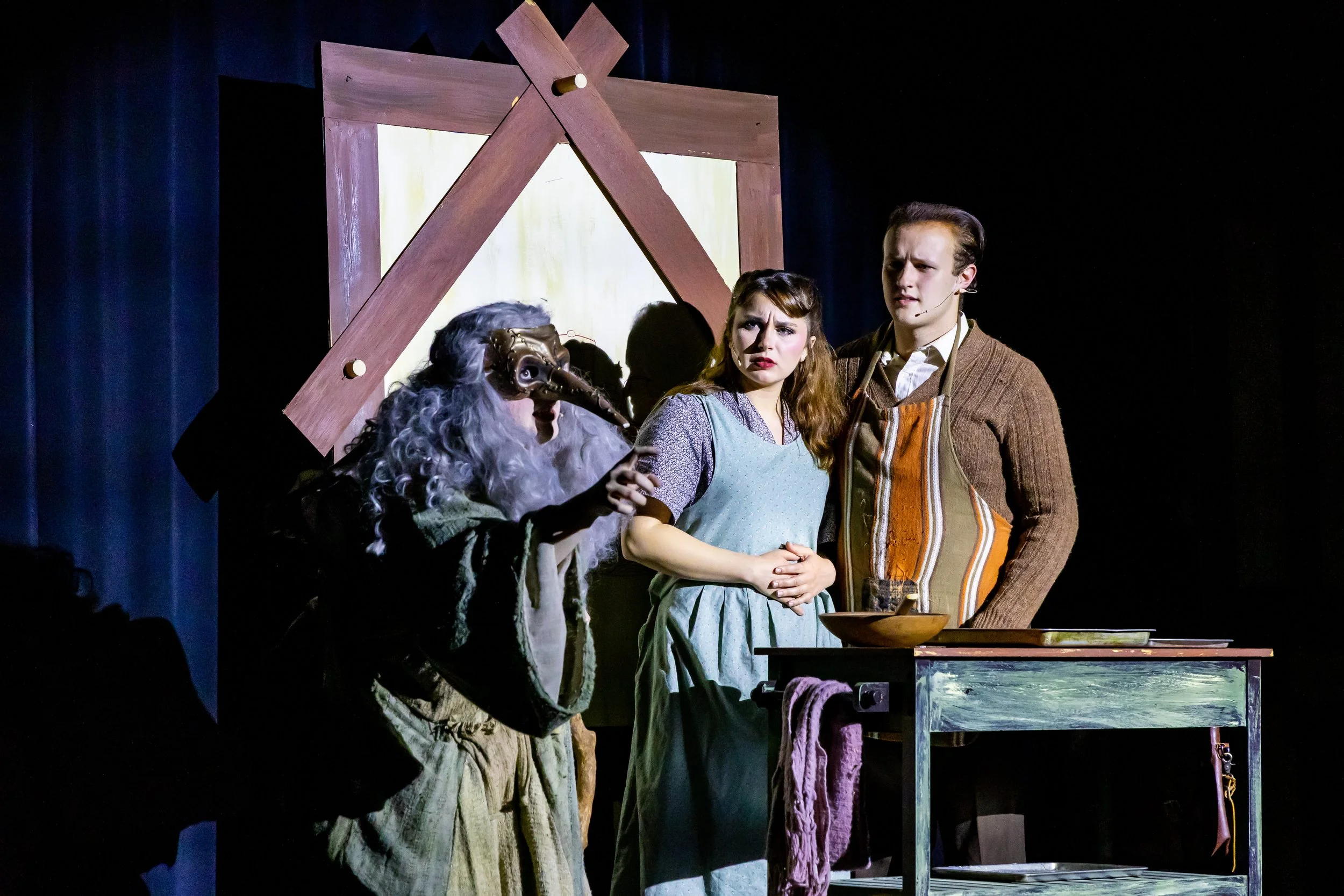 Theater scene with three actors: a woman in a mask with long gray hair, a woman with brown hair in a blue dress, and a man with dark hair in a brown sweater, standing in front of a wooden backdrop resembling a frame.
