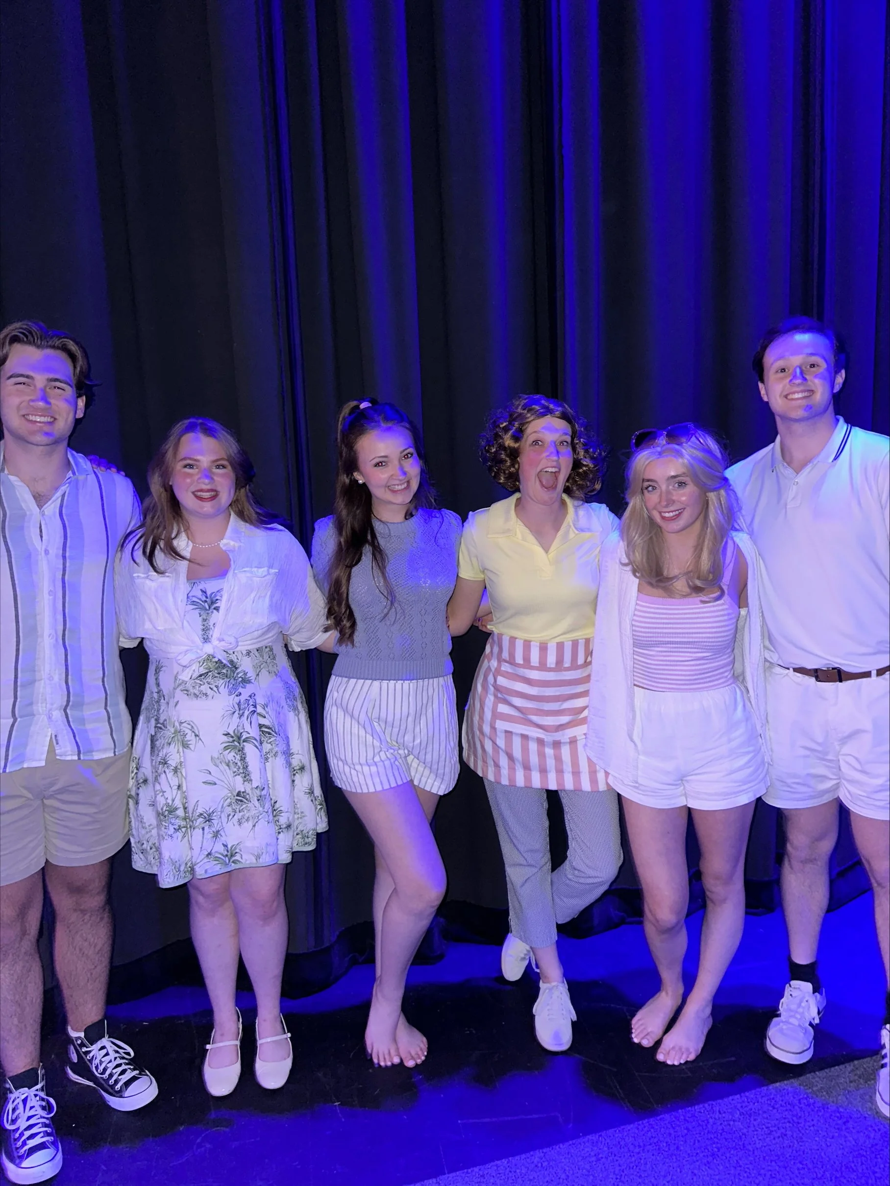 Group of six young adults standing together on stage with dark curtains in the background, smiling and posing for a photo.