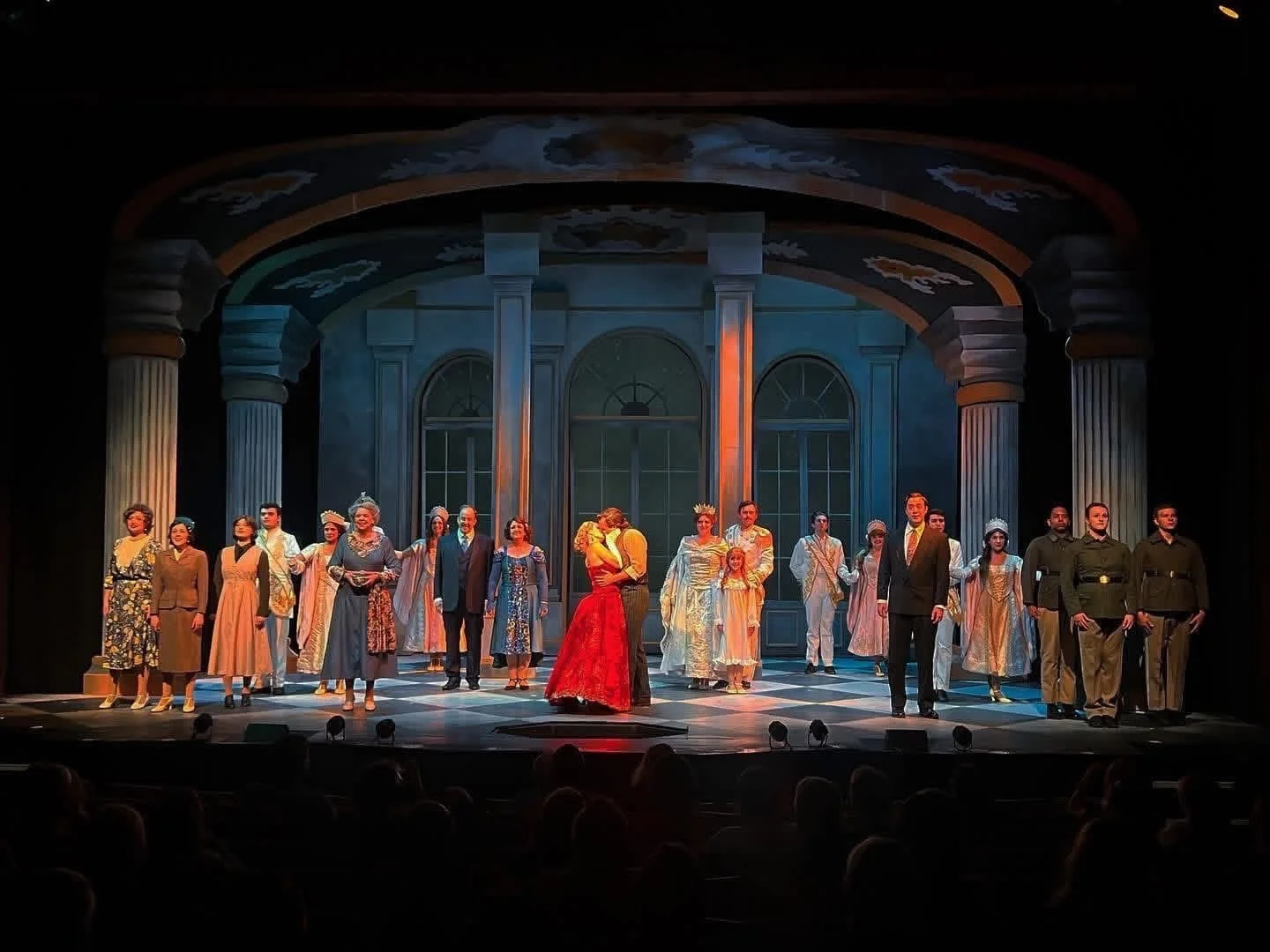 Stage scene from a theatrical performance with actors in period costumes standing in front of a grand set with columns and large windows, some actors embracing or holding hands.