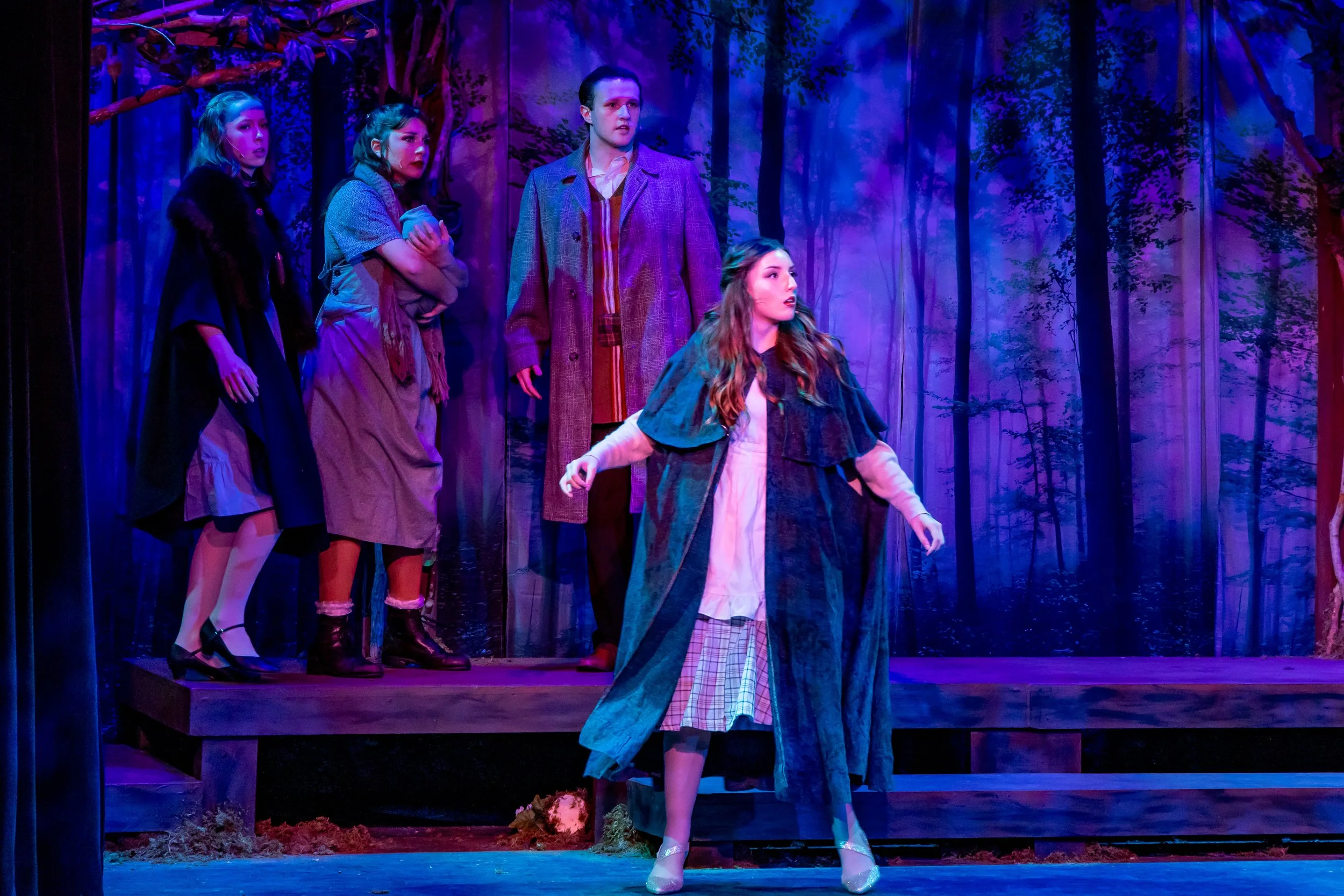 Actors performing on stage in a forest scene, with four women and one man, dressed in vintage clothing, and a woman in front wearing a long coat.