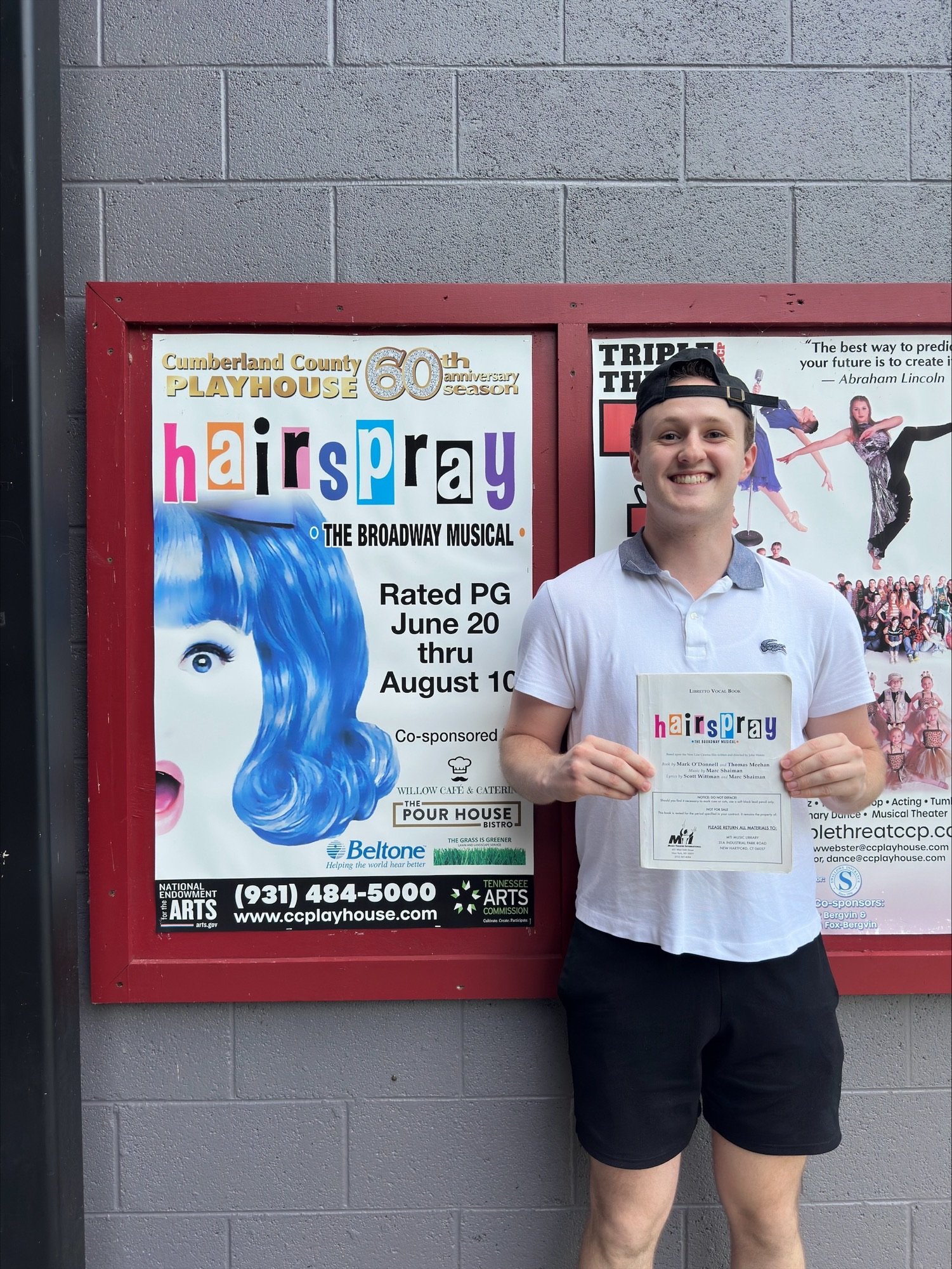 A young man with short hair wearing a white polo shirt, black shorts, and a black cap is smiling while holding a theater ticket and standing in front of a poster for the musical 'Hairspray' at Cumberland County Playhouse.
