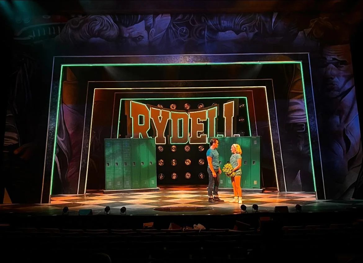 Stage with neon lights spelling RYDELL, two actors in casual clothing, one holding a bouquet of yellow flowers, on a checkered floor, backdrop with lockers and circular lights.