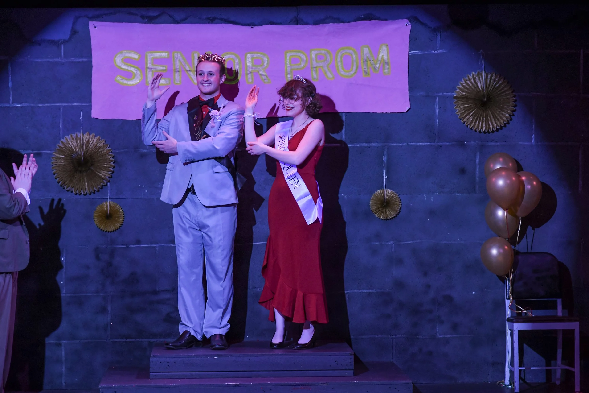 Two young people standing on a stage during a prom, with a man wearing a gray suit and a woman in a red dress with a sash that reads 'Prom Queen'. They are waving to the audience, with balloons and paper decorations in the background and a banner tha