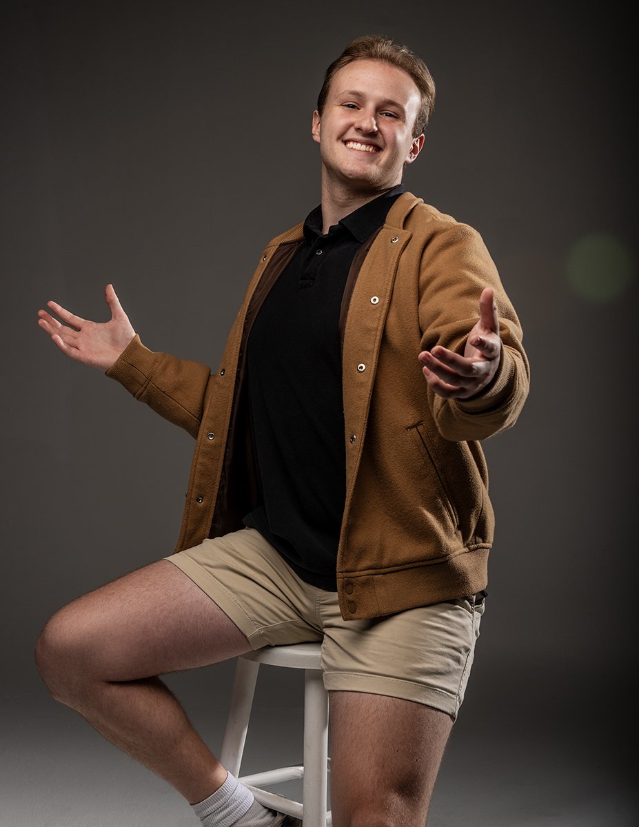 A young man sitting on a white stool, smiling and extending his arms out, wearing a brown jacket, black t-shirt, beige shorts, and white socks, with a plain dark grey background.
