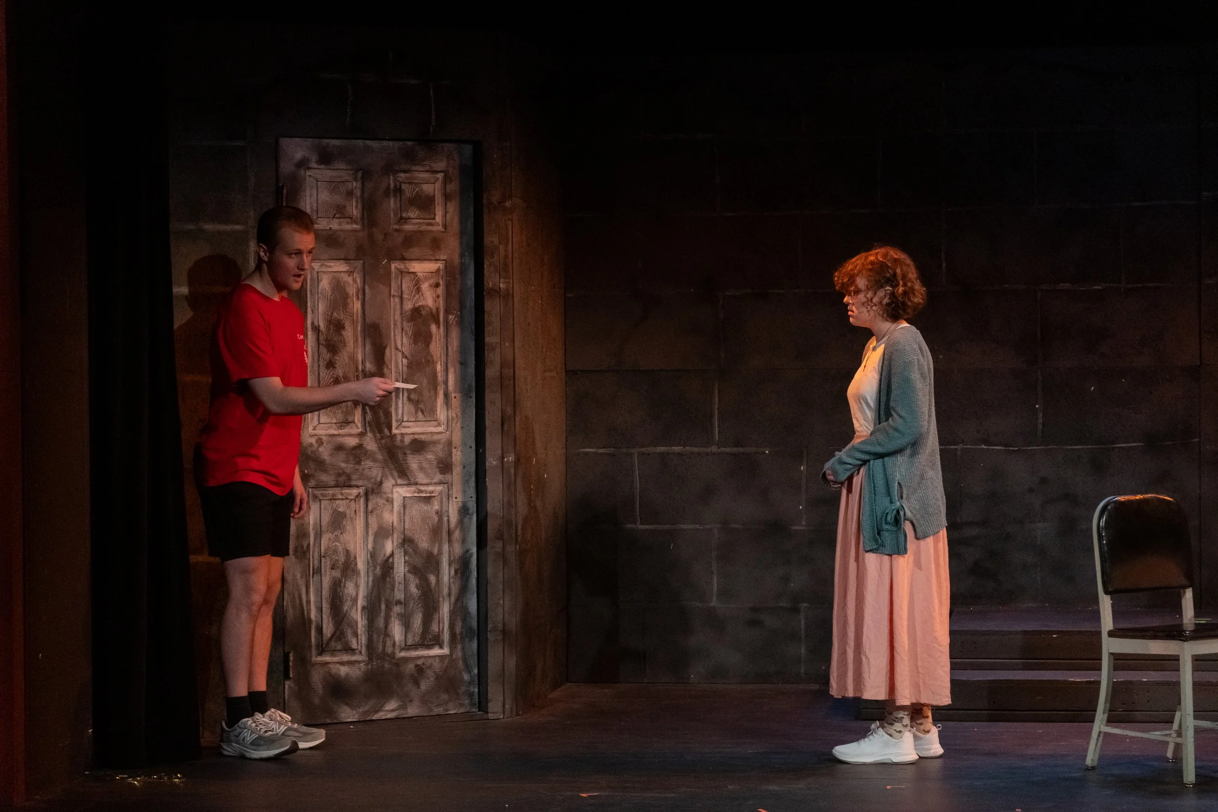 Two women on stage during a theatrical performance, one on the left wearing a red shirt and black shorts holding a paper, the other on the right wearing a gray cardigan and peach skirt, facing each other with a dark wall and a closed door in the back