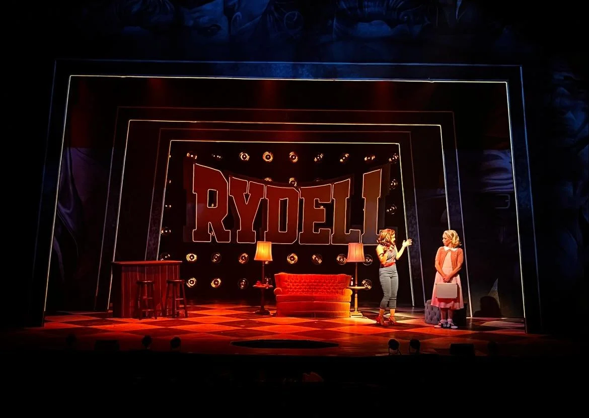 Stage scene from the play 'Rydell', with two women engaged in conversation. The stage is decorated with a red sofa, lamps, and a table, with the play's title 'RYDELL' in large illuminated letters in the background.