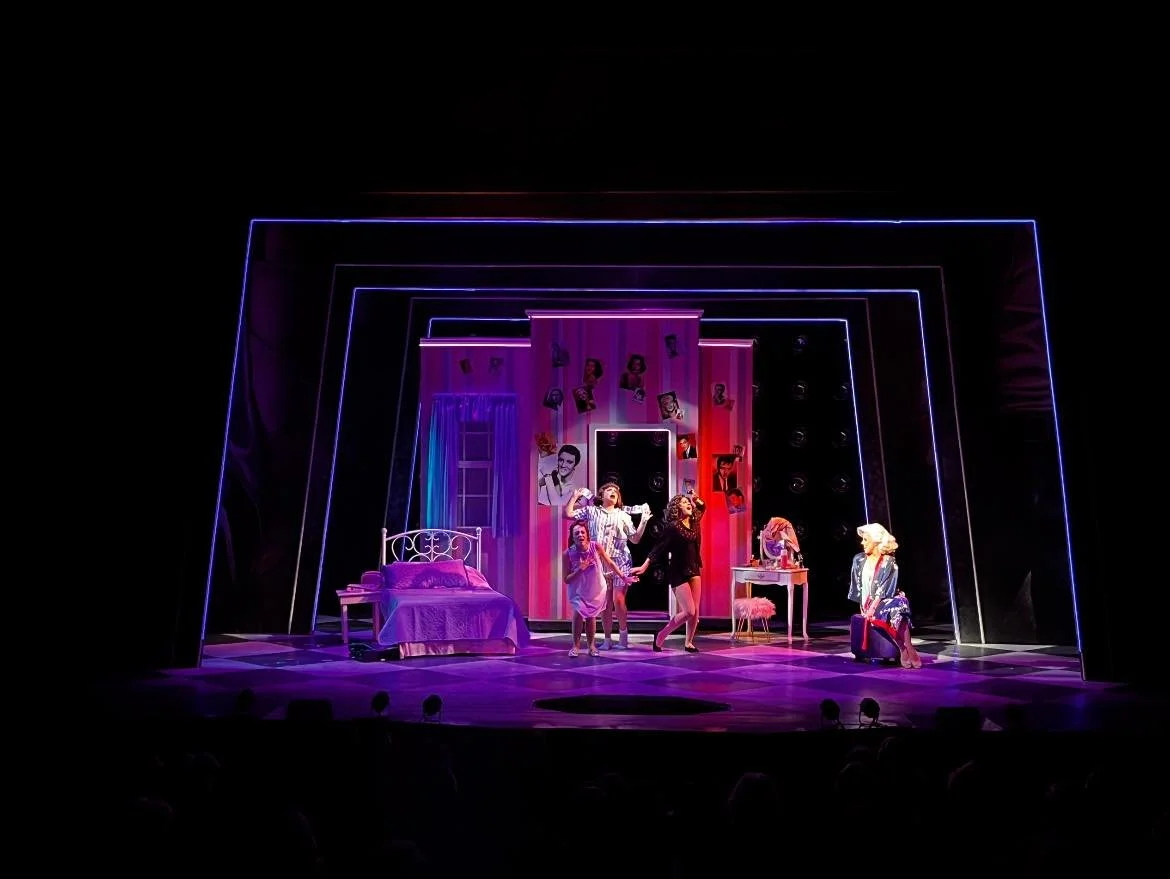 Theatrical stage scene featuring five actors in vintage costumes, with a bedroom set decorated with photographs on the backdrop, pink curtains, and neon light framing the stage.