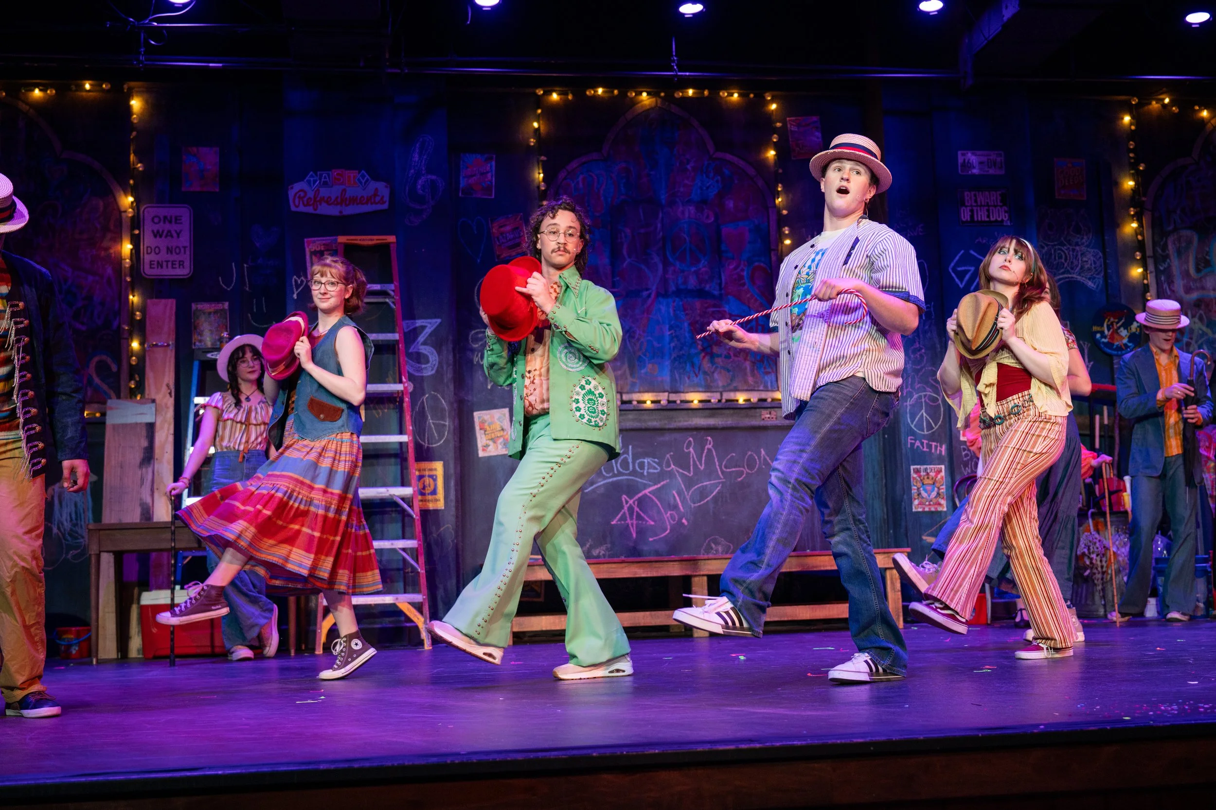 Group of performers in colorful costumes on stage during a theatrical performance, with a graffiti-style backdrop and string lights.