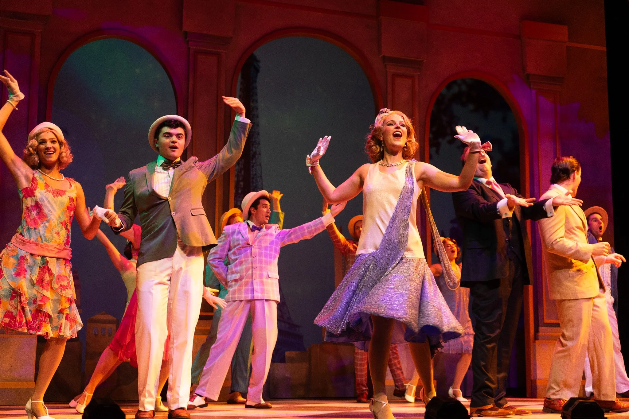 A group of performers on stage during a musical, dressed in colorful vintage costumes, dancing and singing with a set of large arches in the background.
