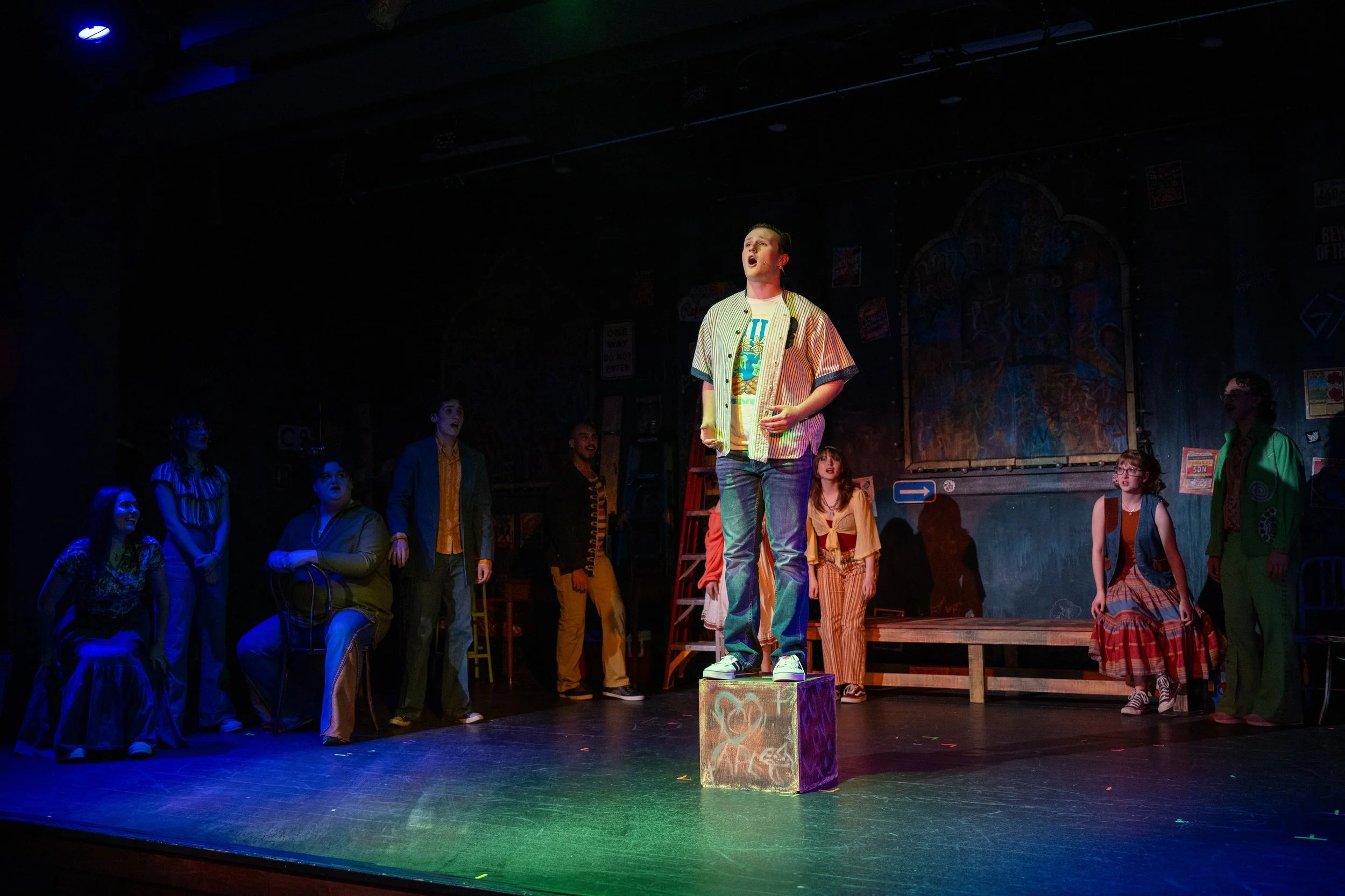 A theatrical stage scene with a young man standing on a colorful box center stage, singing or speaking, surrounded by other actors in casual and vintage clothing, with a dark painted backdrop and theatrical lighting.
