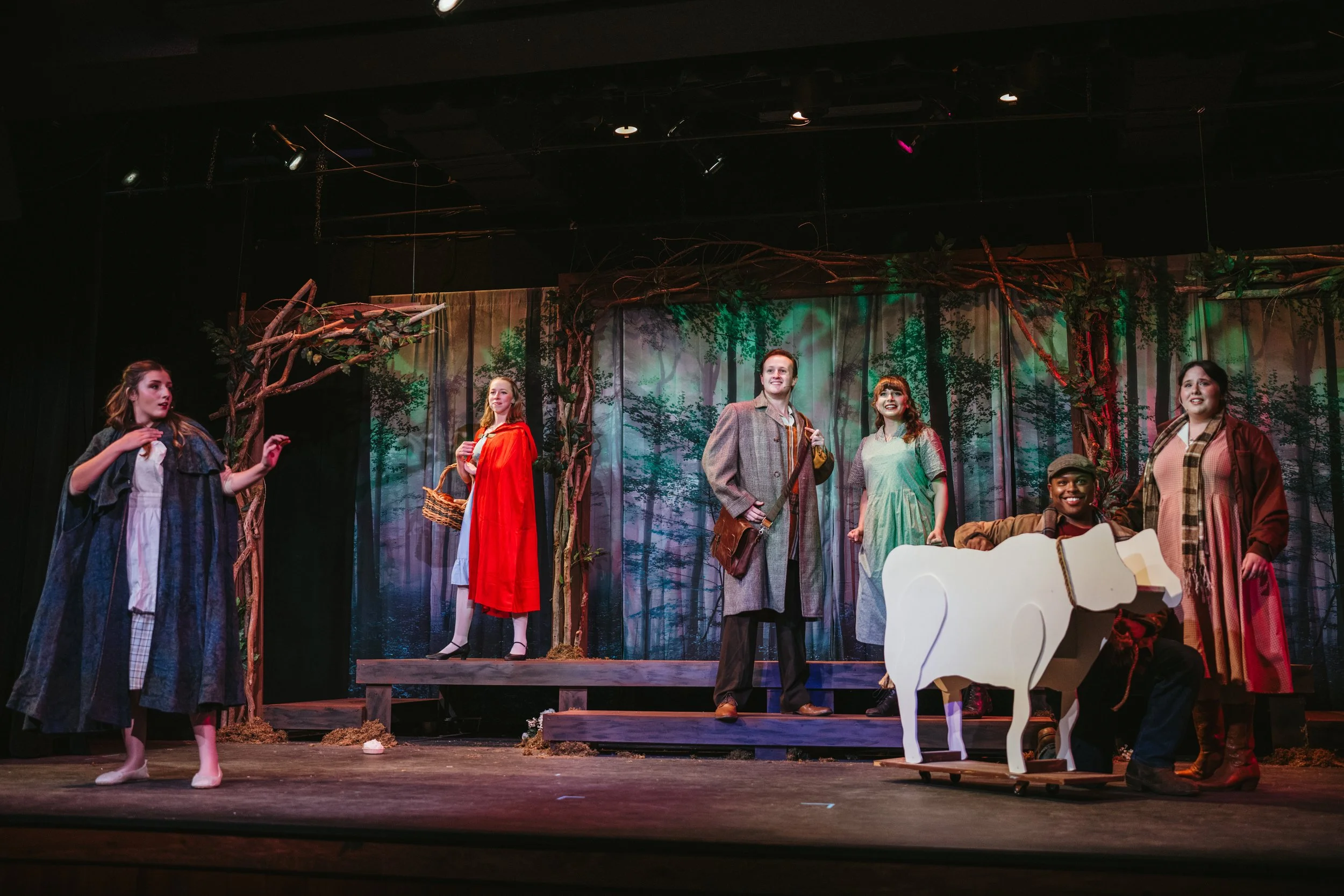 Theater cast performing on stage with forest scenery, actors in costumes, and a white cardboard cow prop.
