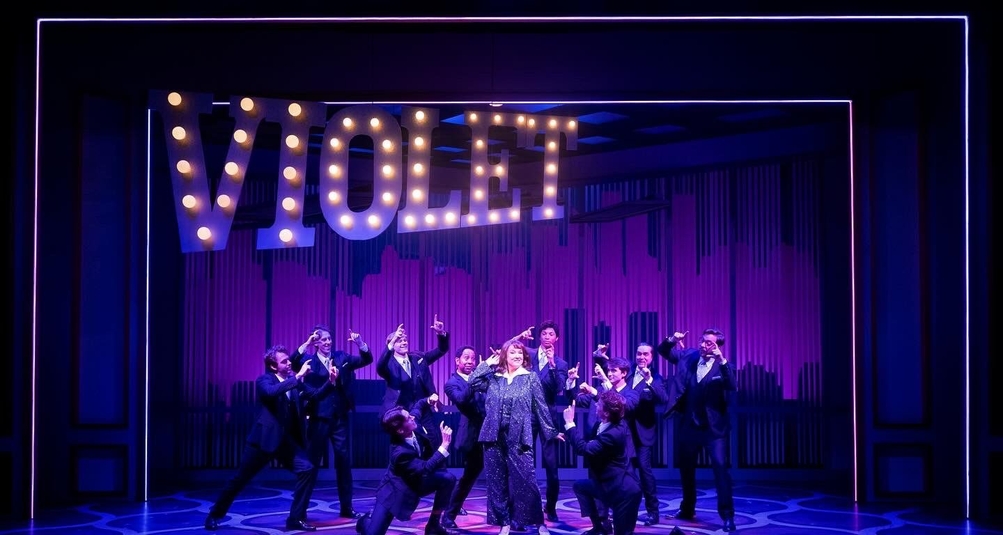 A theatrical stage performance featuring a woman in a sparkly outfit surrounded by ten people dressed in black suits. The stage is decorated with a large illuminated 'VOTE' sign hanging above. The scene is lit with purple lighting.