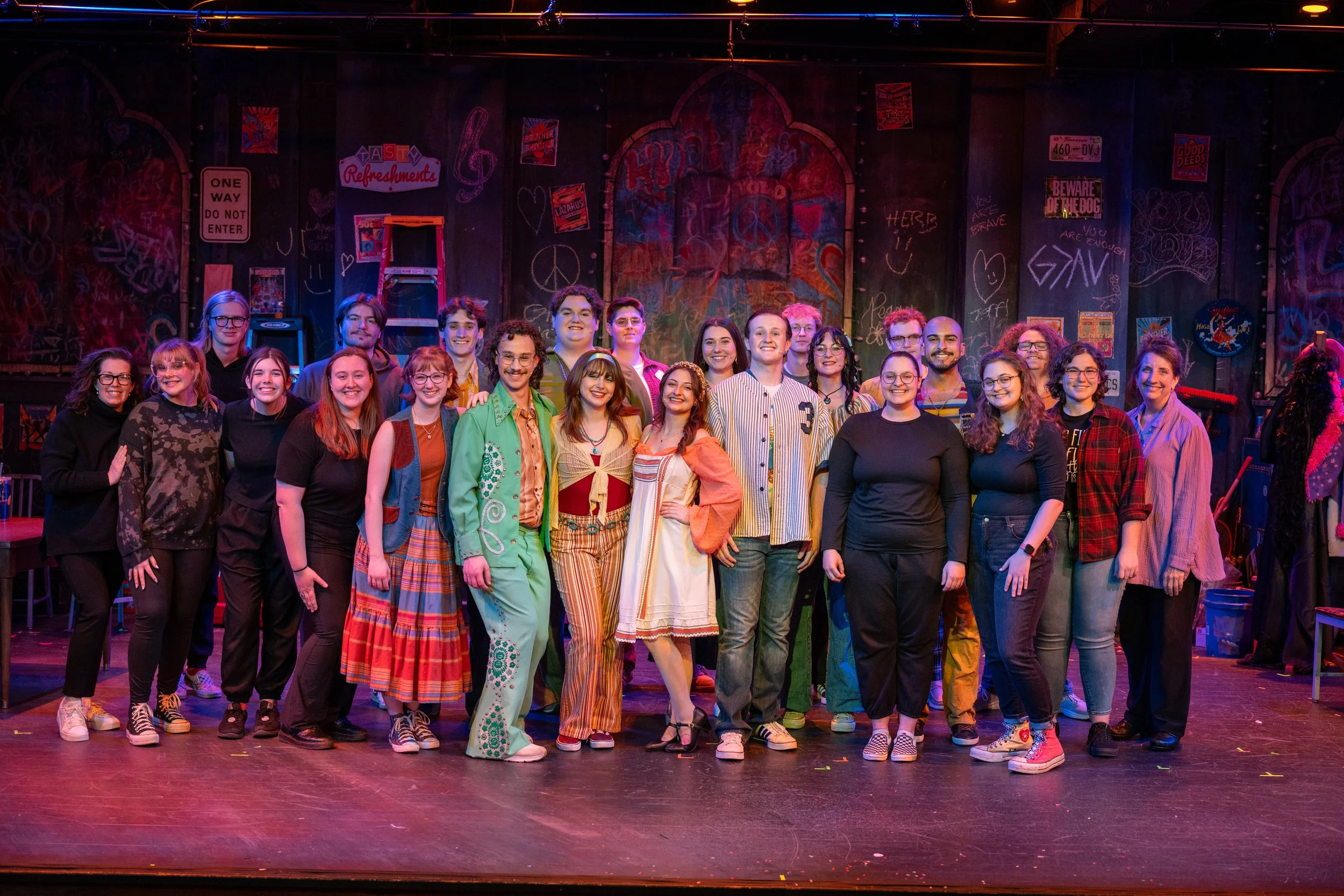 Group of people standing on stage after a performance, colorful graffiti and signs in the background.