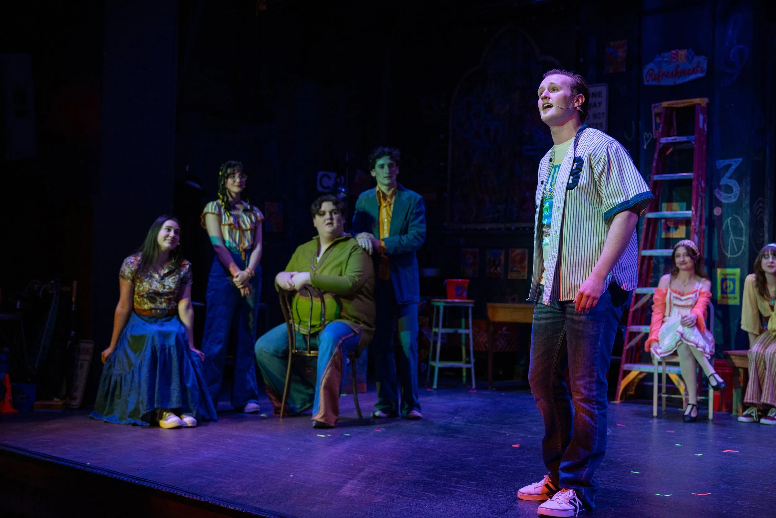 Actors performing a scene on stage, with one male actor standing and singing or speaking, surrounded by six other actors seated or standing, all dressed in colorful costumes, in a theatrical set that resembles a classroom or community center.
