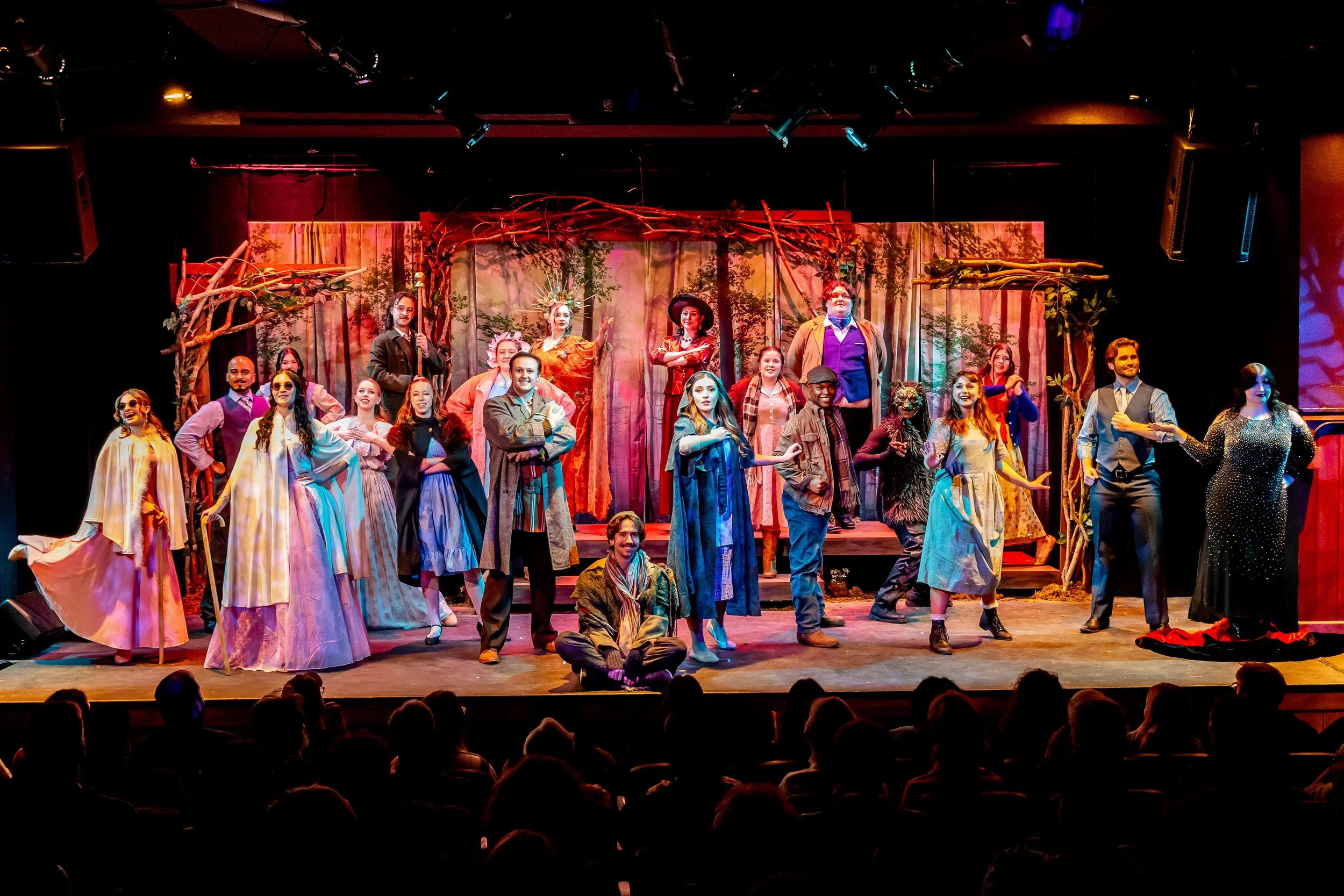 A theatrical stage filled with performers in colorful costumes, standing and sitting in various poses, with a backdrop of trees and branches, and an audience watching from the darkened foreground.