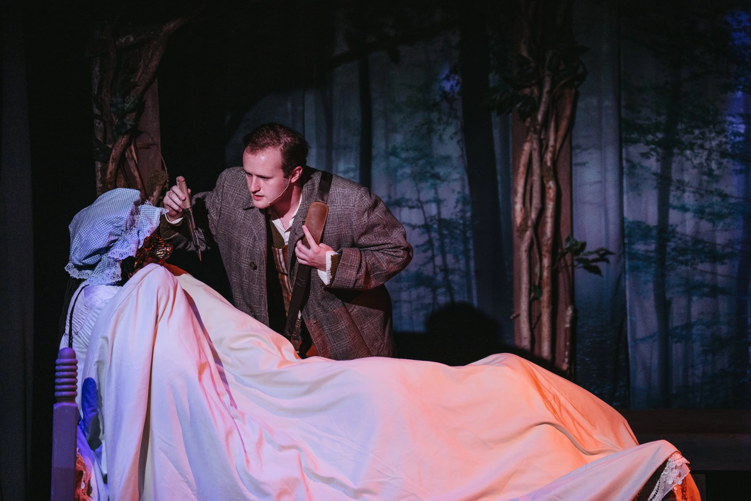 Theater scene of a man in period costume looking intently at a woman lying on a bed in a forest setting background.