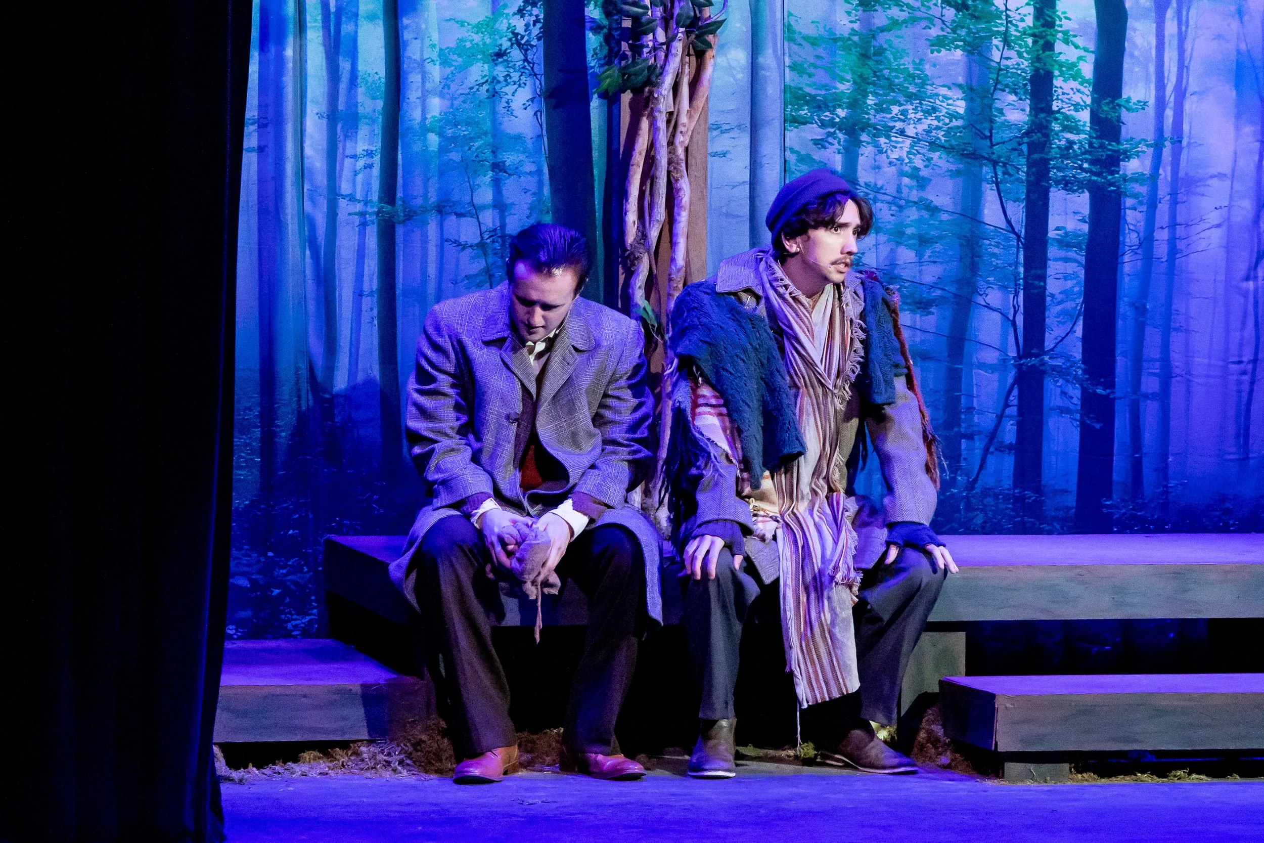 Two actors sitting on a stage with a forest scene background, dressed in old-fashioned costumes, participating in a theatrical performance.