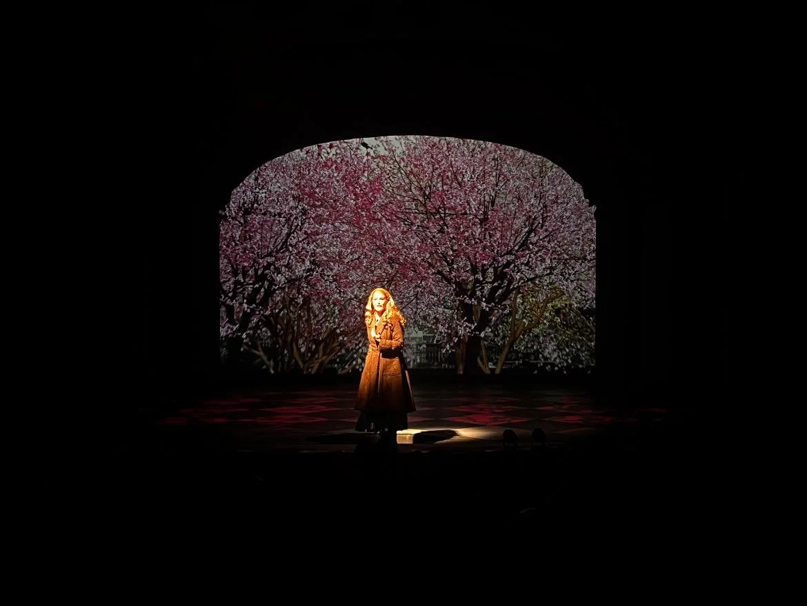 A woman standing on stage in front of a backdrop of blossoming pink and white trees, under a dark arch.