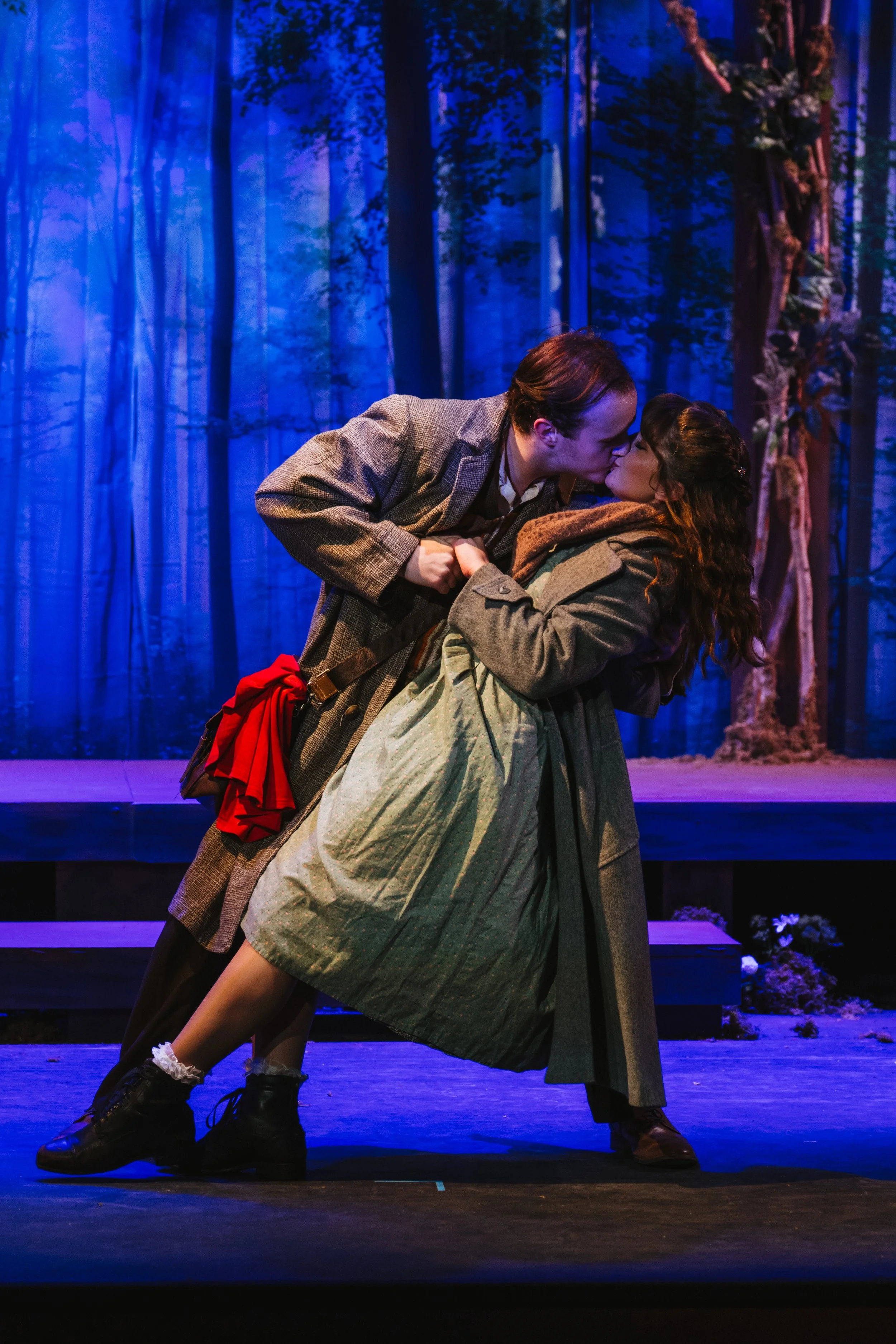 Two actors performing on stage, kissing, with a background of forest scenery.