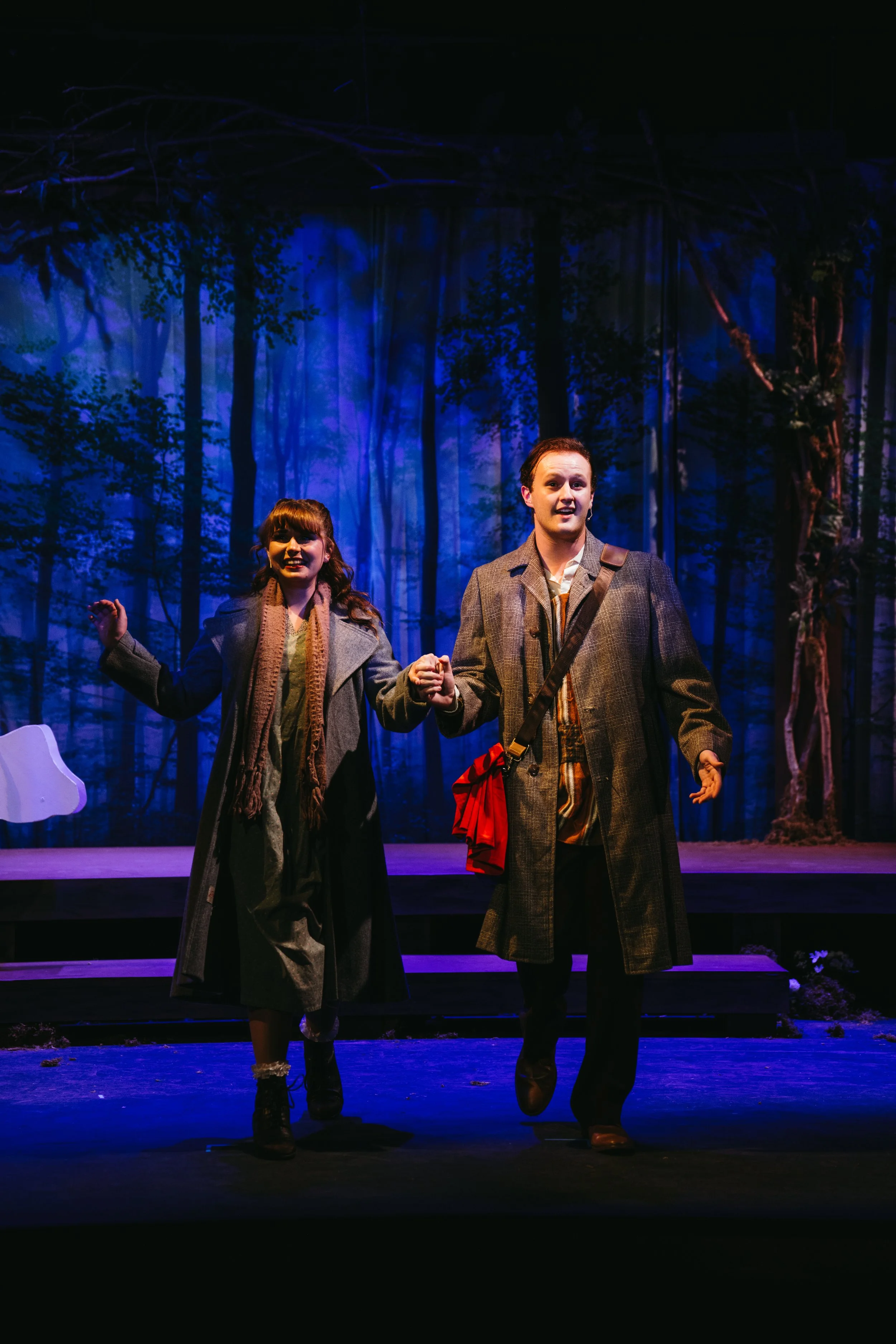 Two actors performing on stage with a forest backdrop, holding hands, dressed in vintage clothing.