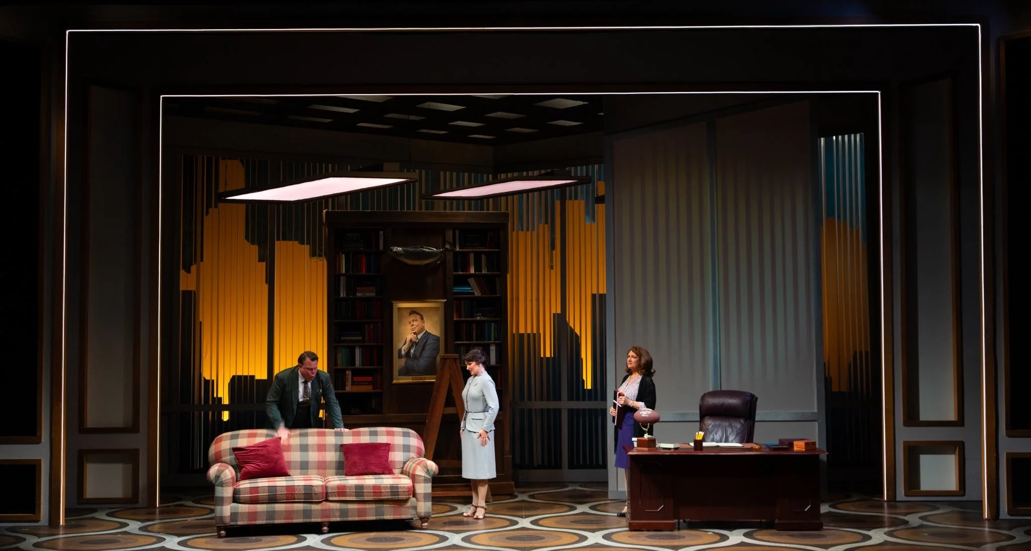 Stage scene from a play depicting a vintage office with three actors, a sofa with red pillows, a desk, a large bookshelf, a portrait of a man, and decorative lighting.