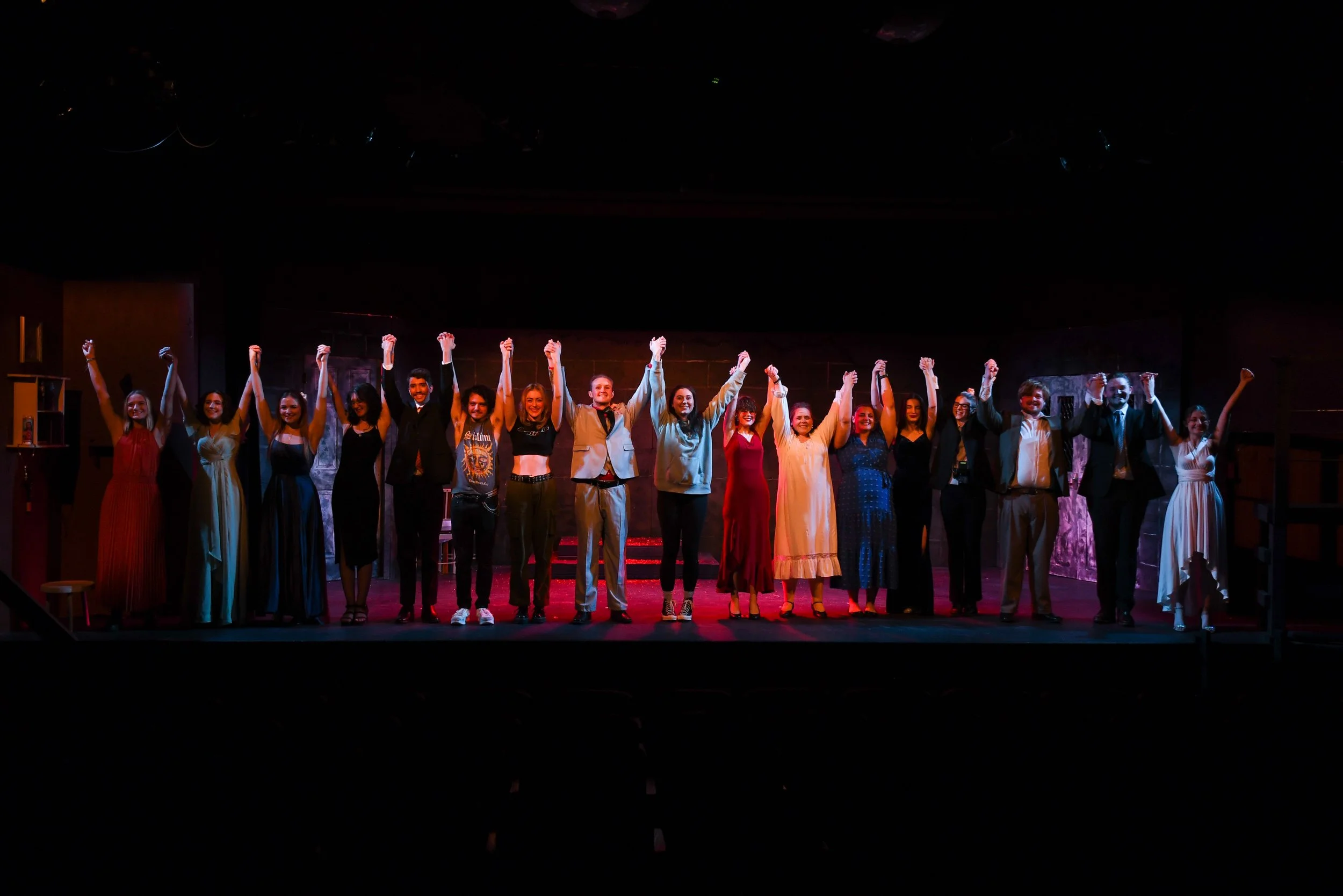 Group of performers standing on stage, holding hands and raising arms in celebration during a theater performance, with stage lighting highlighting them.