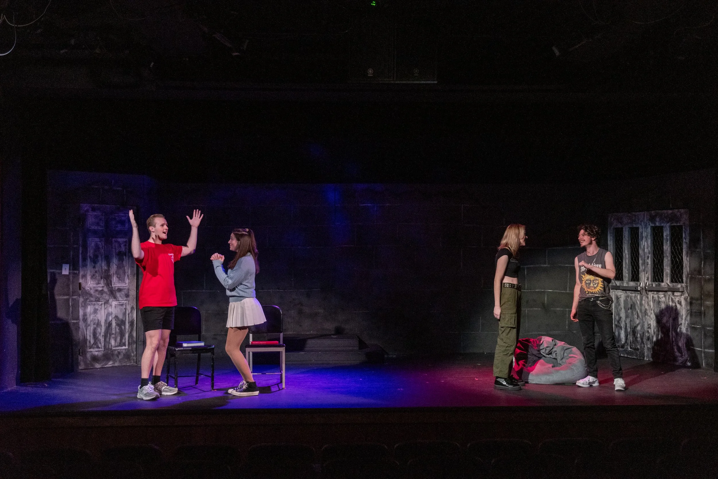 Four young actors on stage during a play, with two people on the left engaging in a conversation and two others on the right talking, part of a theatrical production.