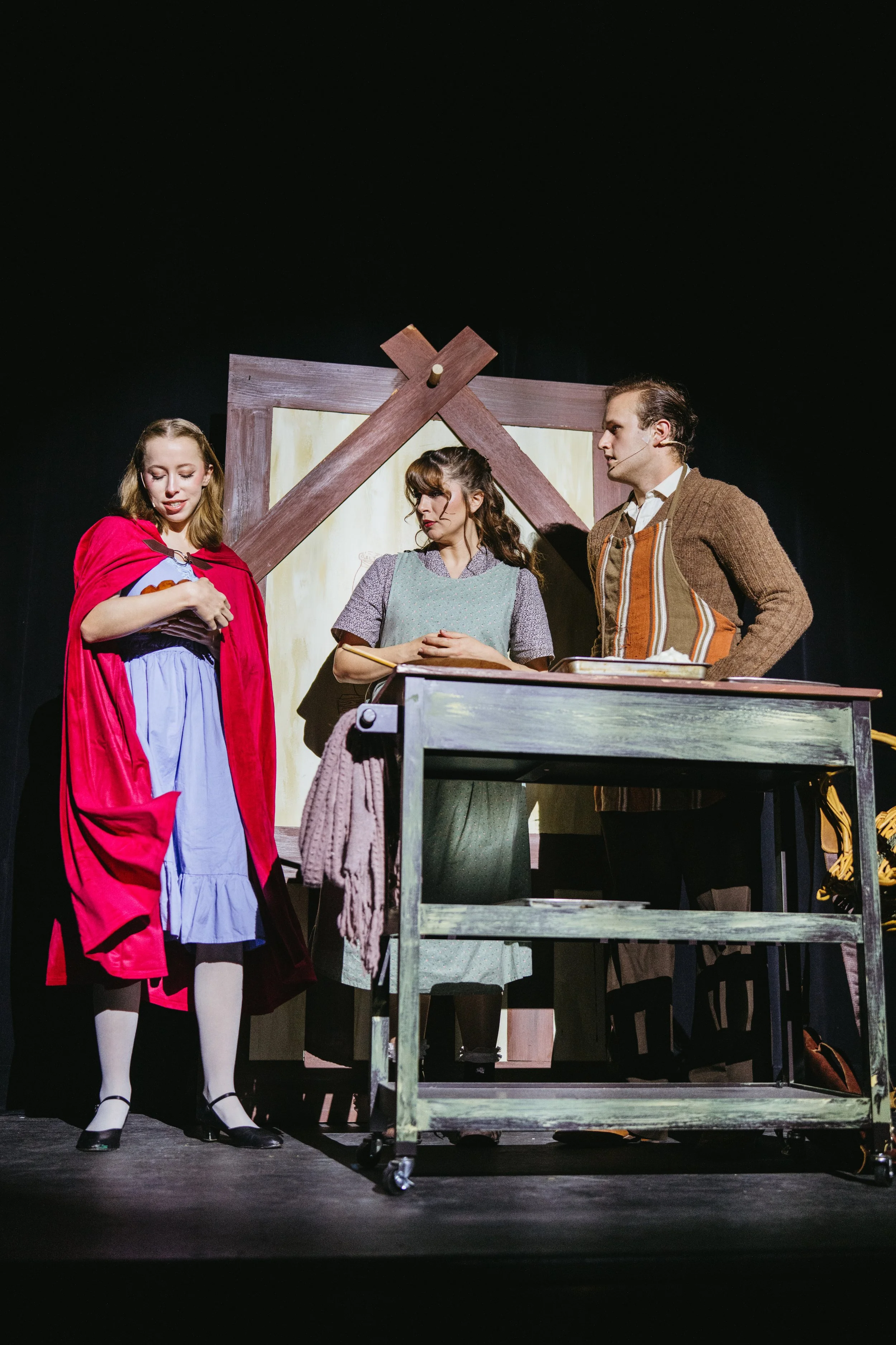 Three actors on stage performing a play set in a historical period, with a wooden structure in the background and a table in front of them. The woman on the left is dressed in a red cloak and vintage dress, while the other two are dressed in period-a