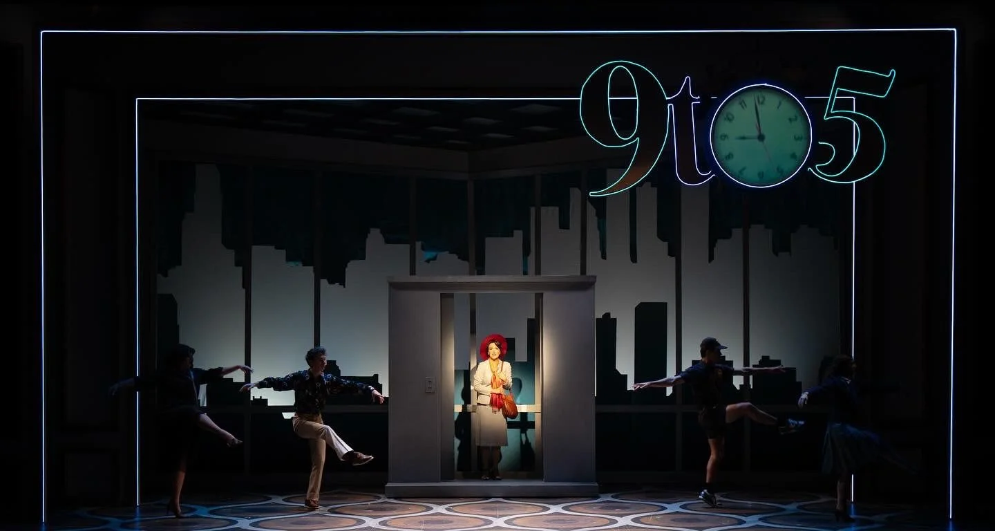 Stage scene with a woman in a red wig and beige coat standing in a small elevator, surrounded by four dancers in a choreographed pose, under a neon sign that reads 'glo5' with an upside-down clock dial.