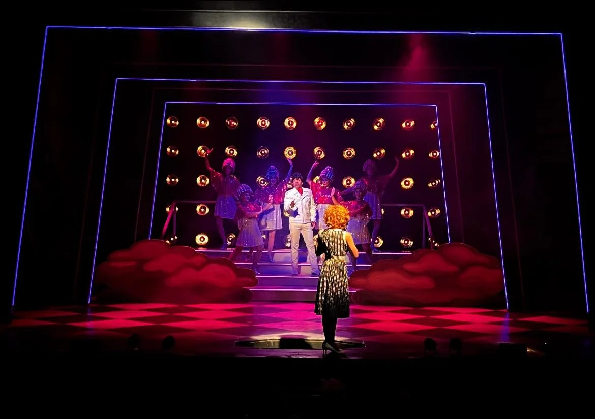 Stage scene from a theatrical play featuring a performer with curly blonde hair in a polka dot dress facing characters on stairs, illuminated with vibrant pink, purple, and yellow lights.