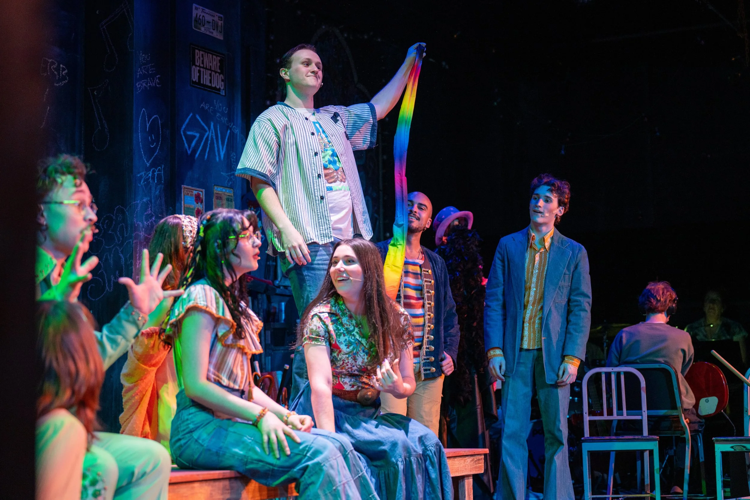 A group of young actors on stage during a theatrical performance, with one person holding a rainbow-colored ribbon aloft, surrounded by colorful lighting and set decorations.