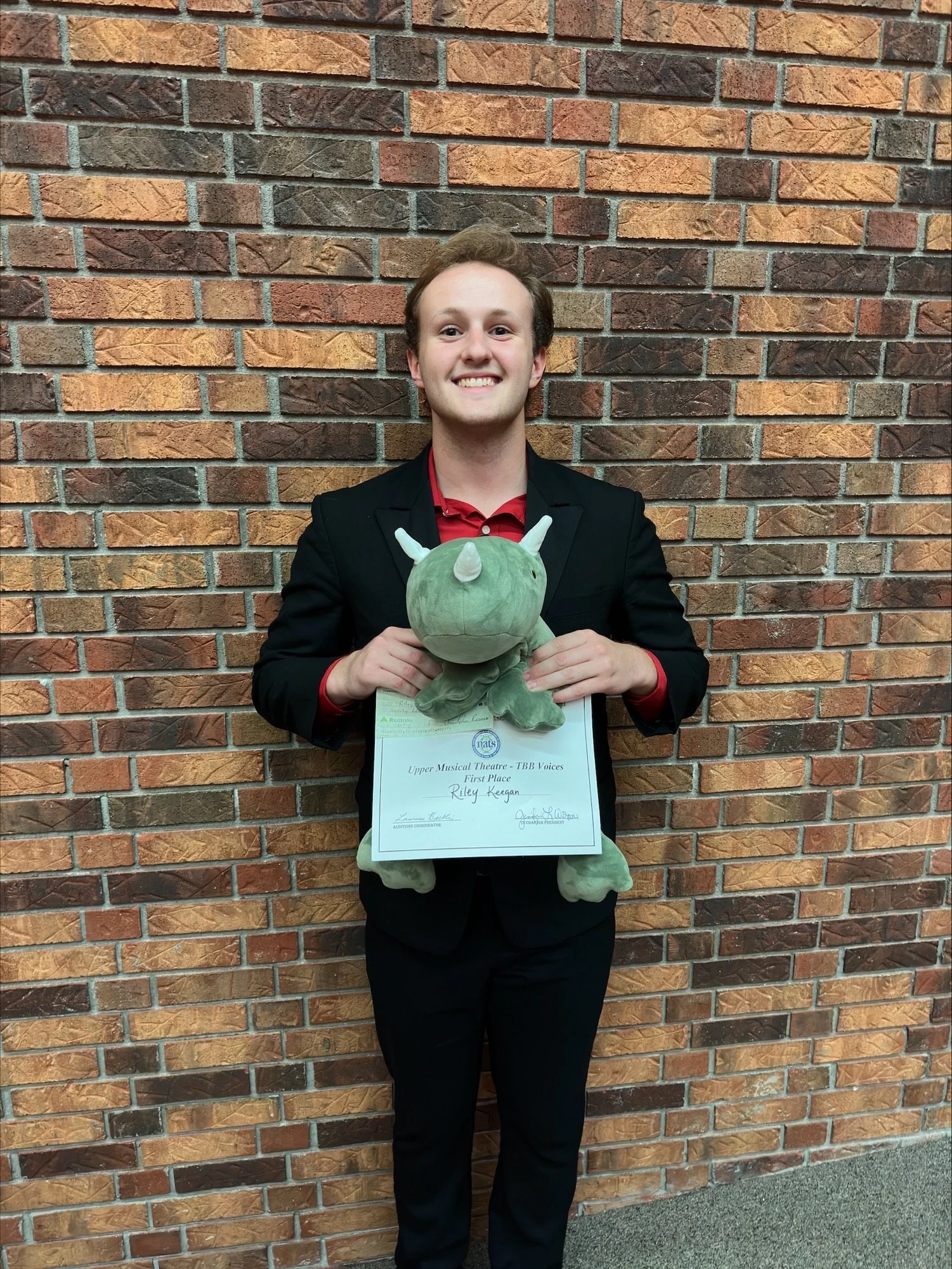 A young man in a black suit and red shirt is standing in front of a brick wall, holding a plush green dinosaur toy and a certificate of achievement for first place in a musical theater competition.