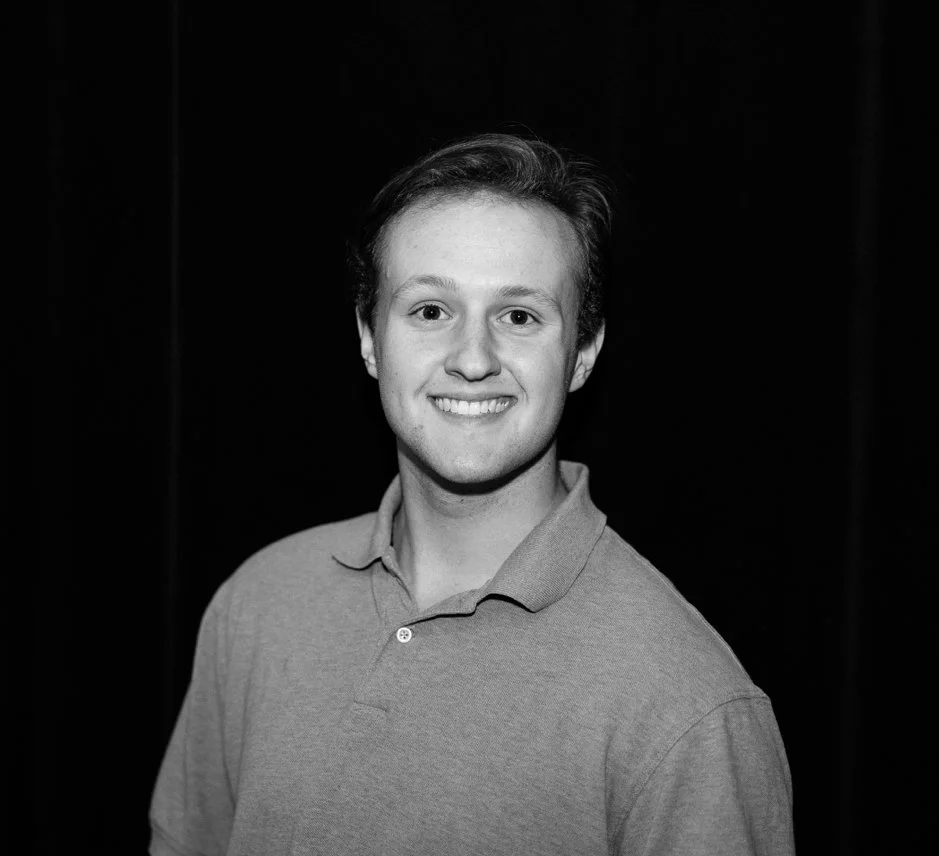 A young man with light skin and short hair smiling, wearing a polo shirt, against a dark background.