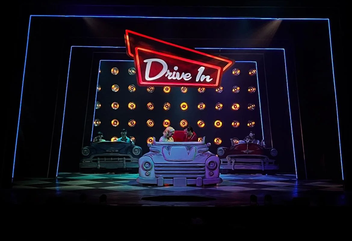 Colorful neon sign reading 'Drive In' with classic car stage set and four people, illuminated with neon lights and circular lights in the background.
