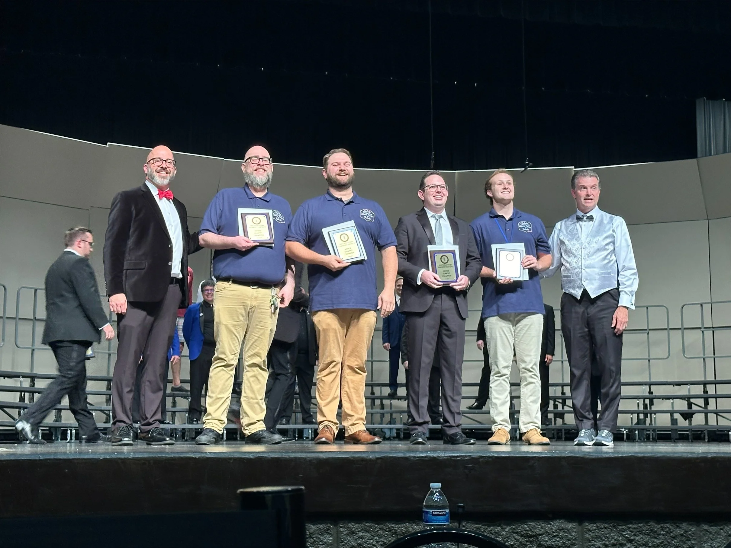 Group of seven men standing on stage, some holding plaques, at an awards ceremony or recognition event in an auditorium.