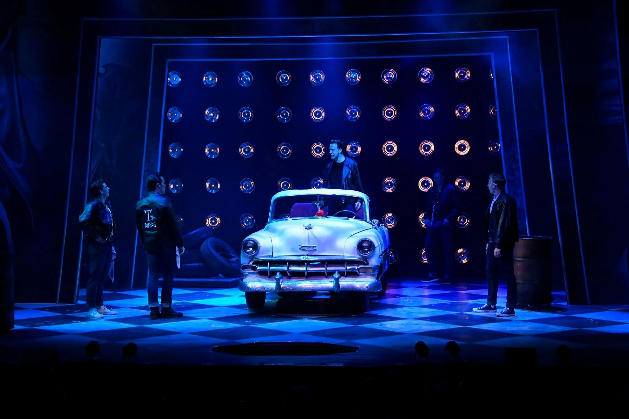 Stage scene with a vintage car and five people, surrounded by neon blue lights and a dark backdrop, resembling a theatrical performance or play.