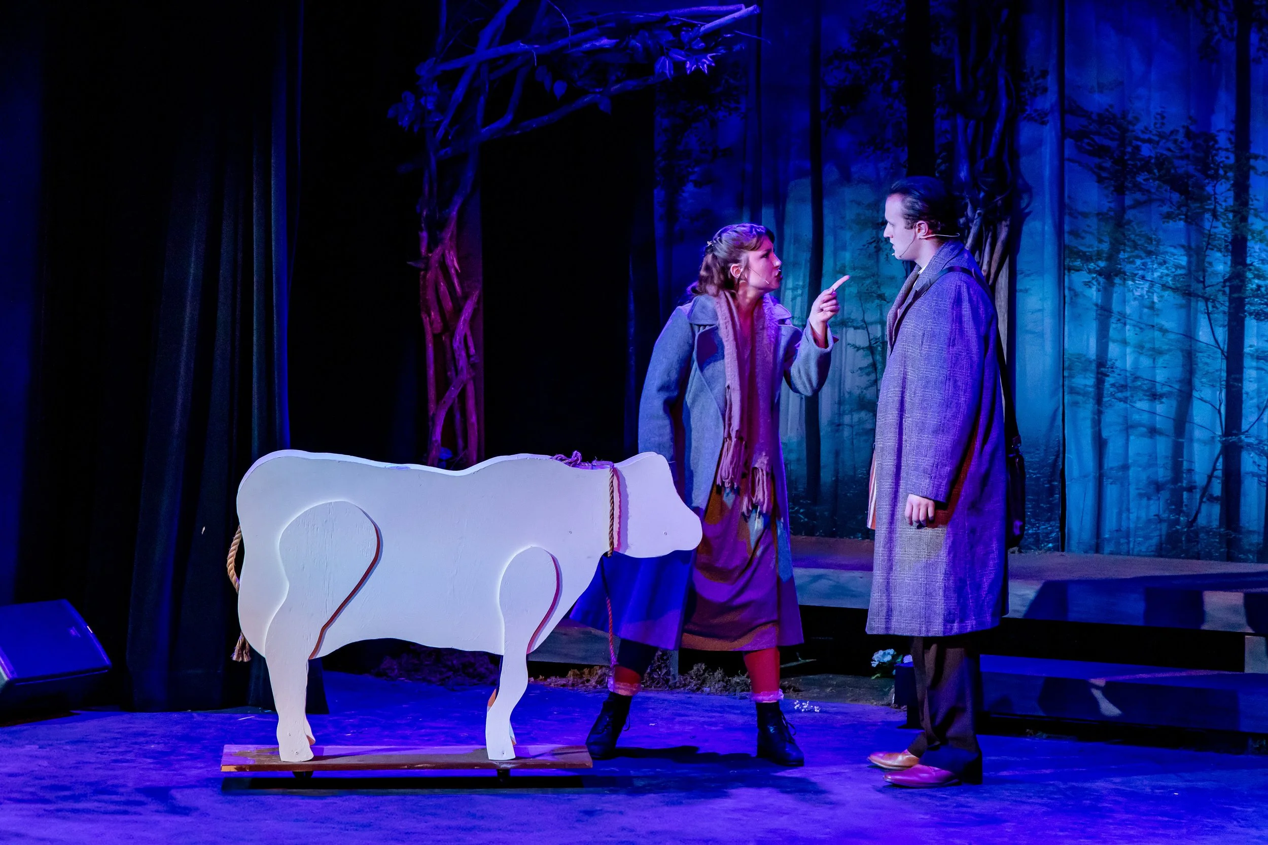 Two actors perform on a stage with a forest background and a white, wooden animal prop. The woman is pointing a finger at the man, who is standing and listening.