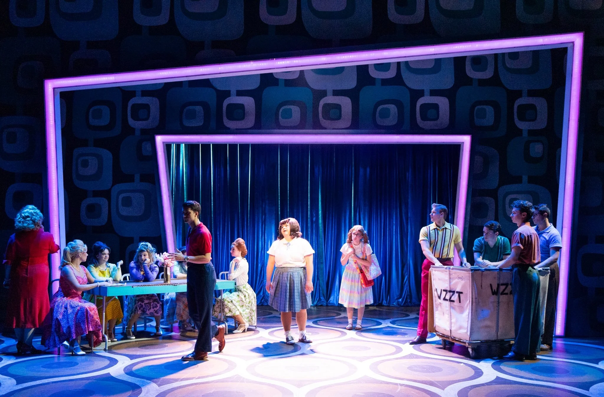 People performing on a colorful stage with geometric patterns and neon lights, set for a theatrical production.