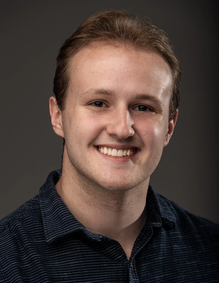 A young man with light brown hair and blue eyes smiling in front of a dark gray background.