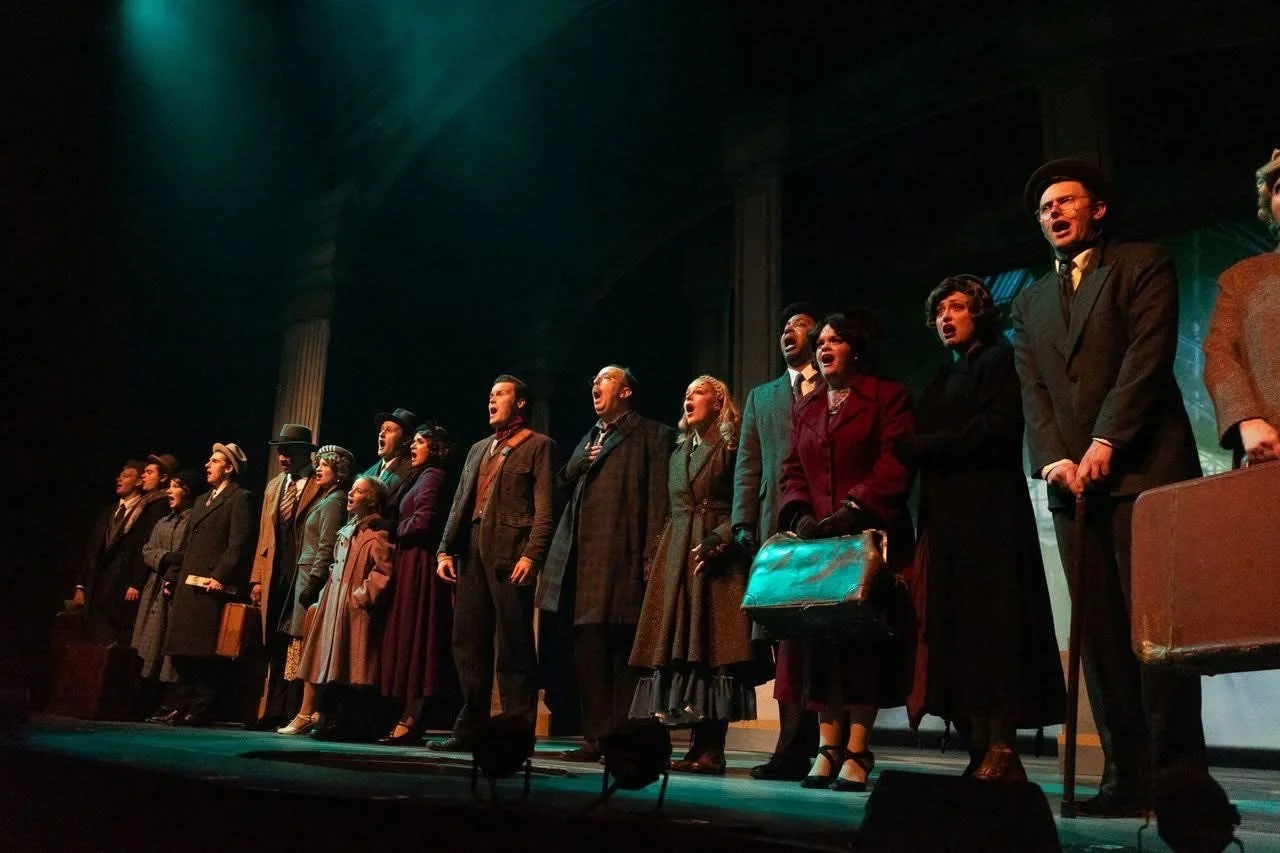The cast of a theater performance standing on stage, dressed in vintage clothing, singing or reciting lines.