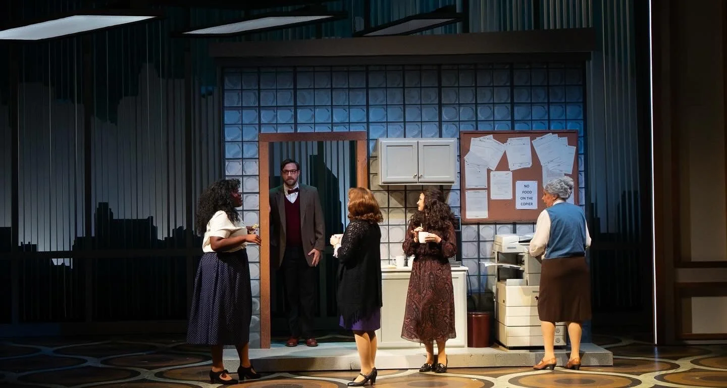 A stage scene with five actors in period costumes, set in an office or waiting room with a bulletin board, desk, and cubicle wall.